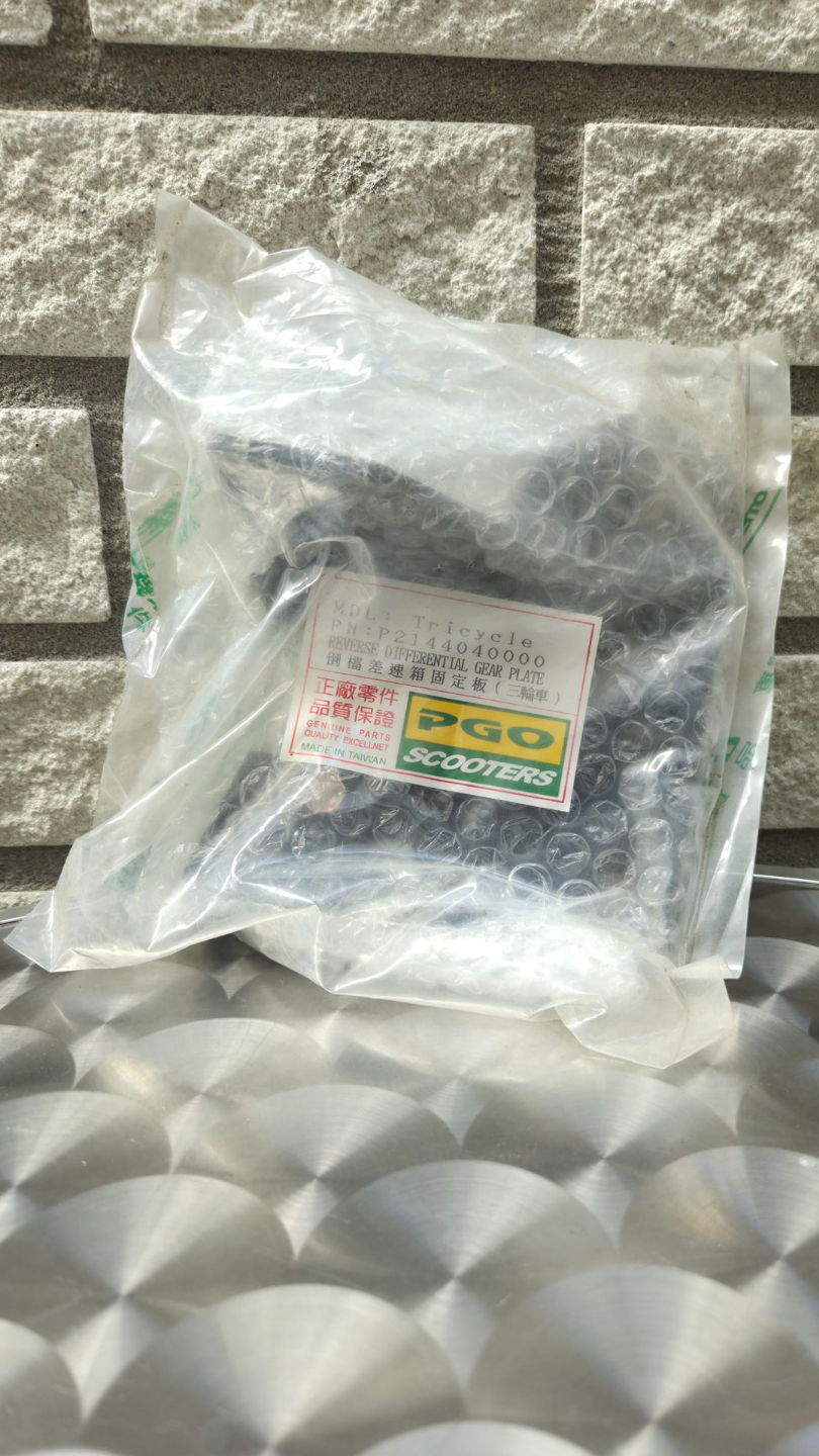 In plastic verpakt zwart voorwerp met etiket, leunend tegen een stenen muur op een zilverkleurige, gestructureerde ondergrond.