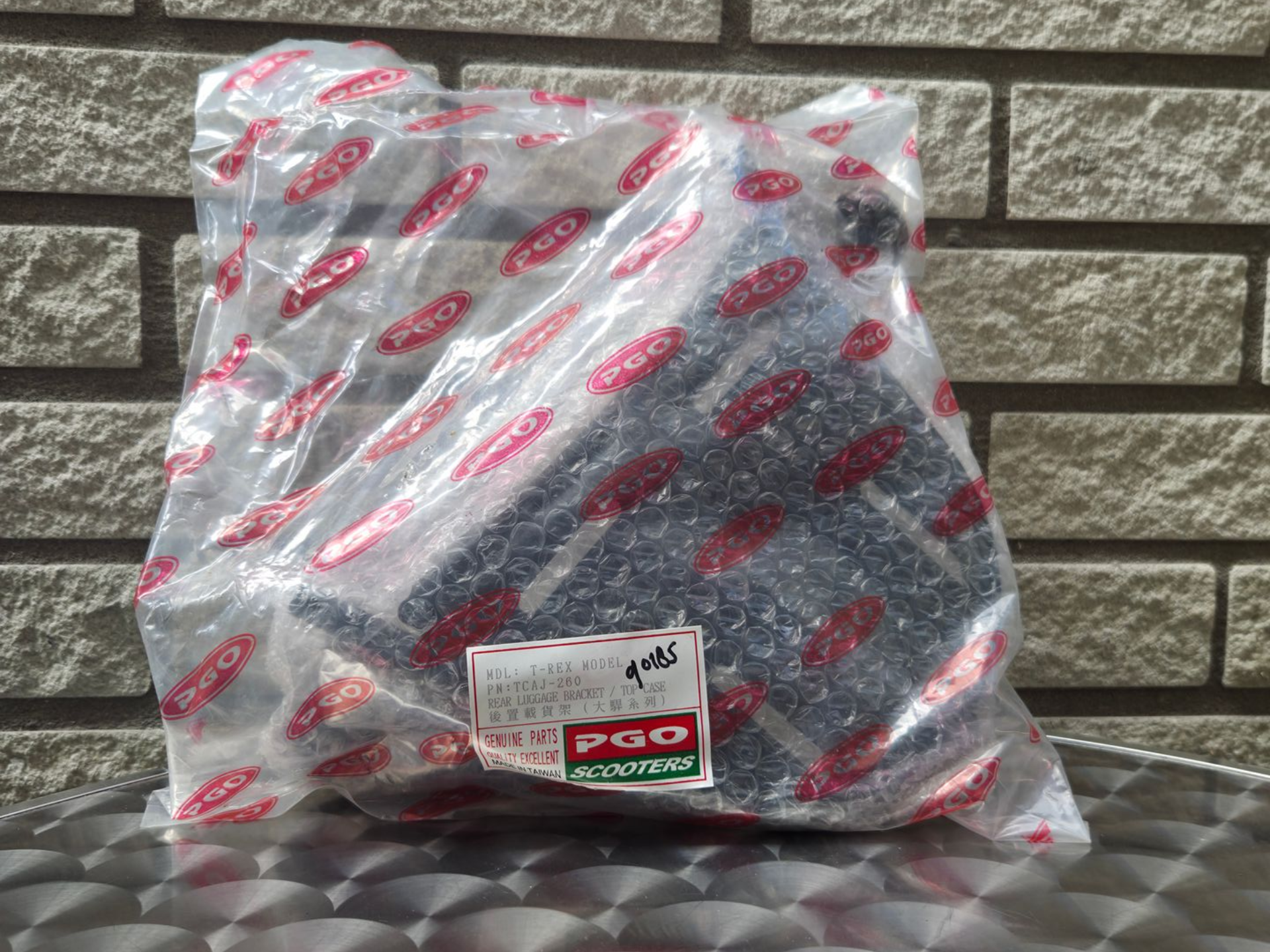 Een doorzichtige plastic zak met rood-zwarte onderdelen en een wit etiket, die tegen een bakstenen muur leunt.