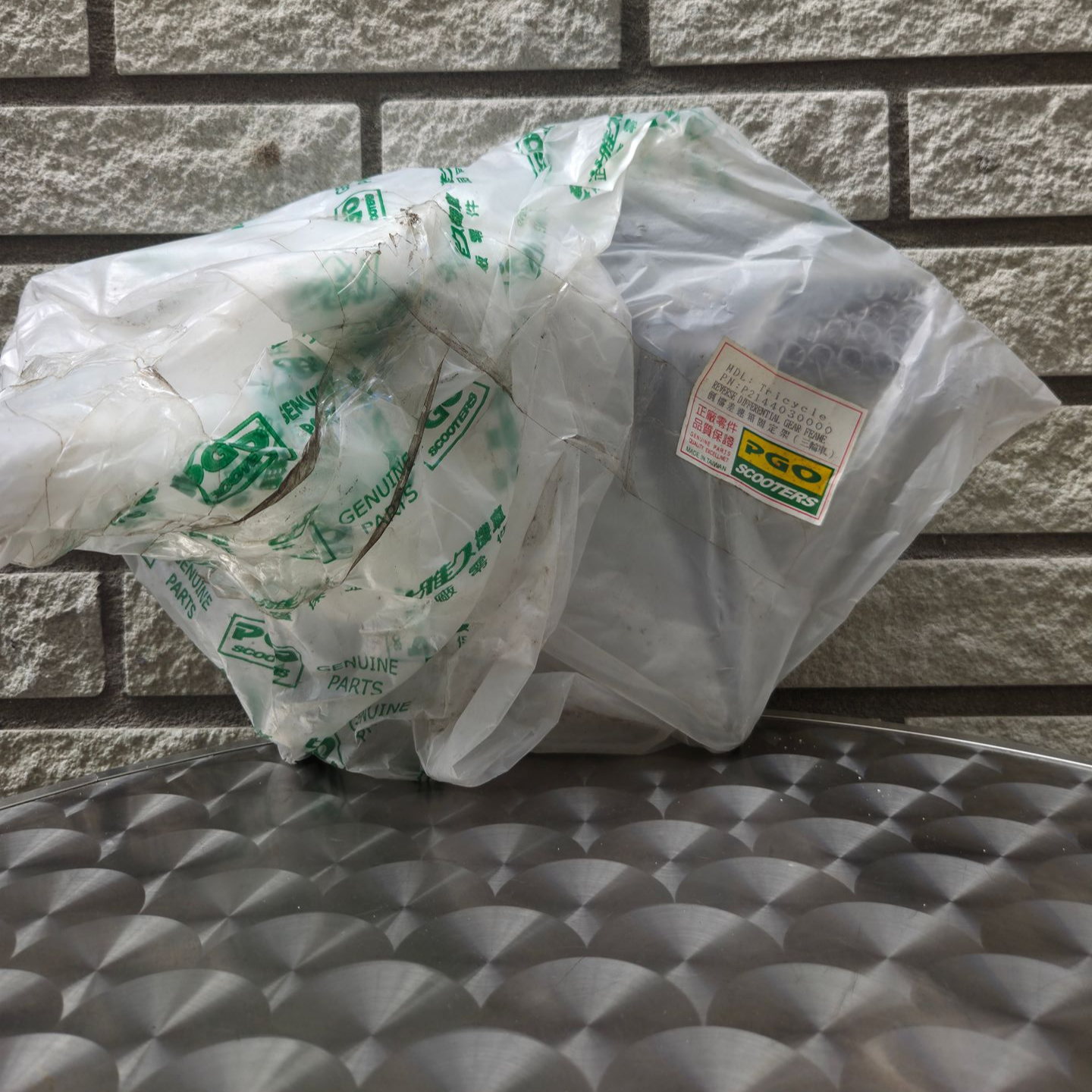 Een plastic zak en een grijze container met een etiket staan ​​op een metalen ondergrond met structuur, tegen een bakstenen muur.
