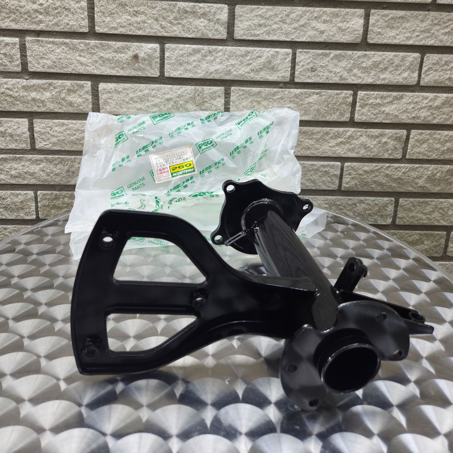 Zwart plastic auto-onderdeel op een oppervlak met patroon, met daarachter een transparante zak en een bakstenen muur op de achtergrond.