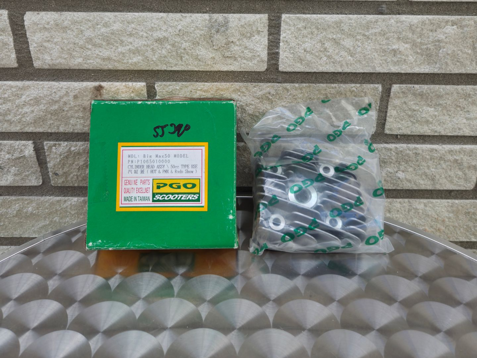 Groene productdoos en ingepakte hardware-onderdelen op een metalen ondergrond met patroon tegen een bakstenen muur.