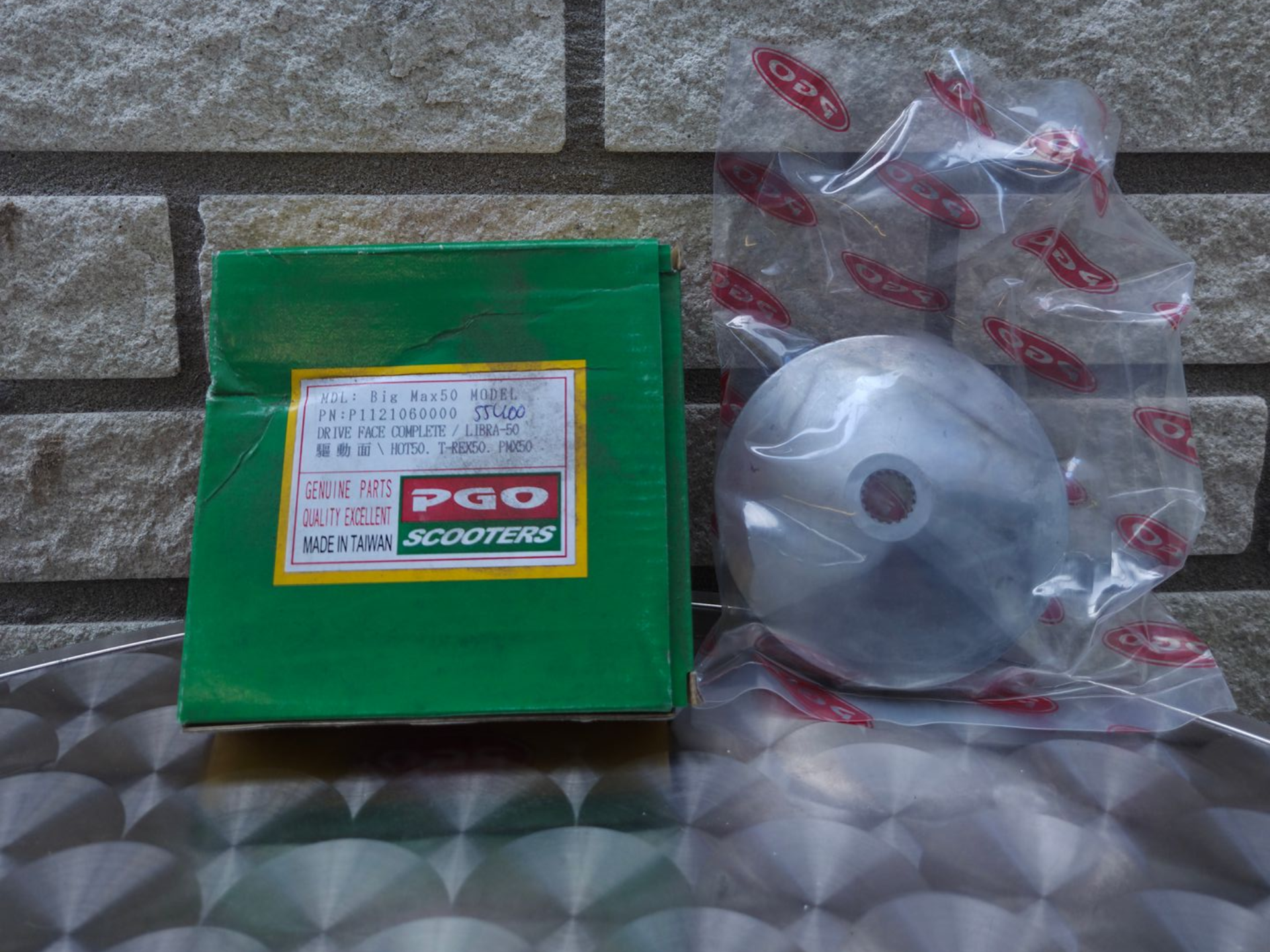 Groene doos met etiket naast een zilverkleurig rond onderdeel, verpakt in een doorzichtige plastic zak op een tegeloppervlak.