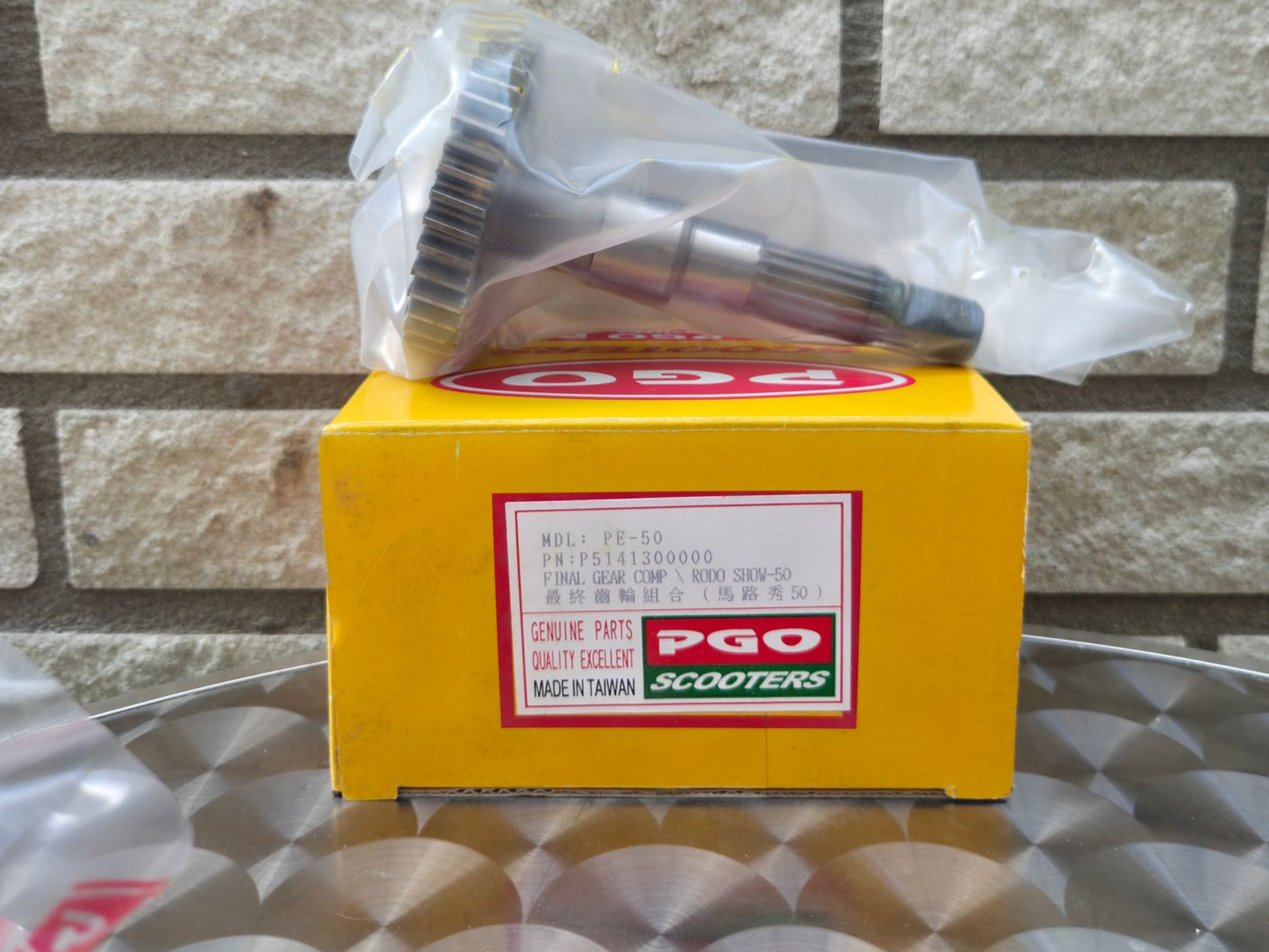 Gele PGO-doos met een onderdelenlabel, met daarop een doorzichtige zak met metalen schroeven op een tafel met een patroon.