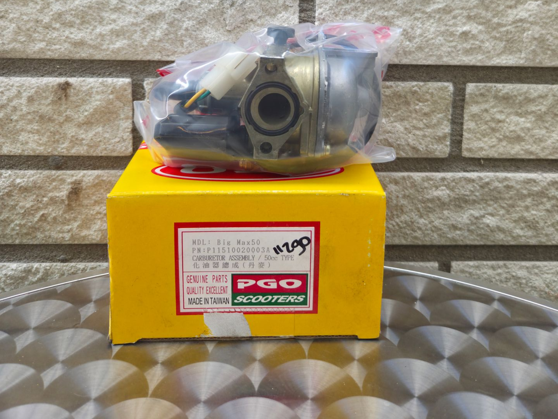 Carburateur verpakt in plastic op een gele doos, met een stenen muur op de achtergrond.