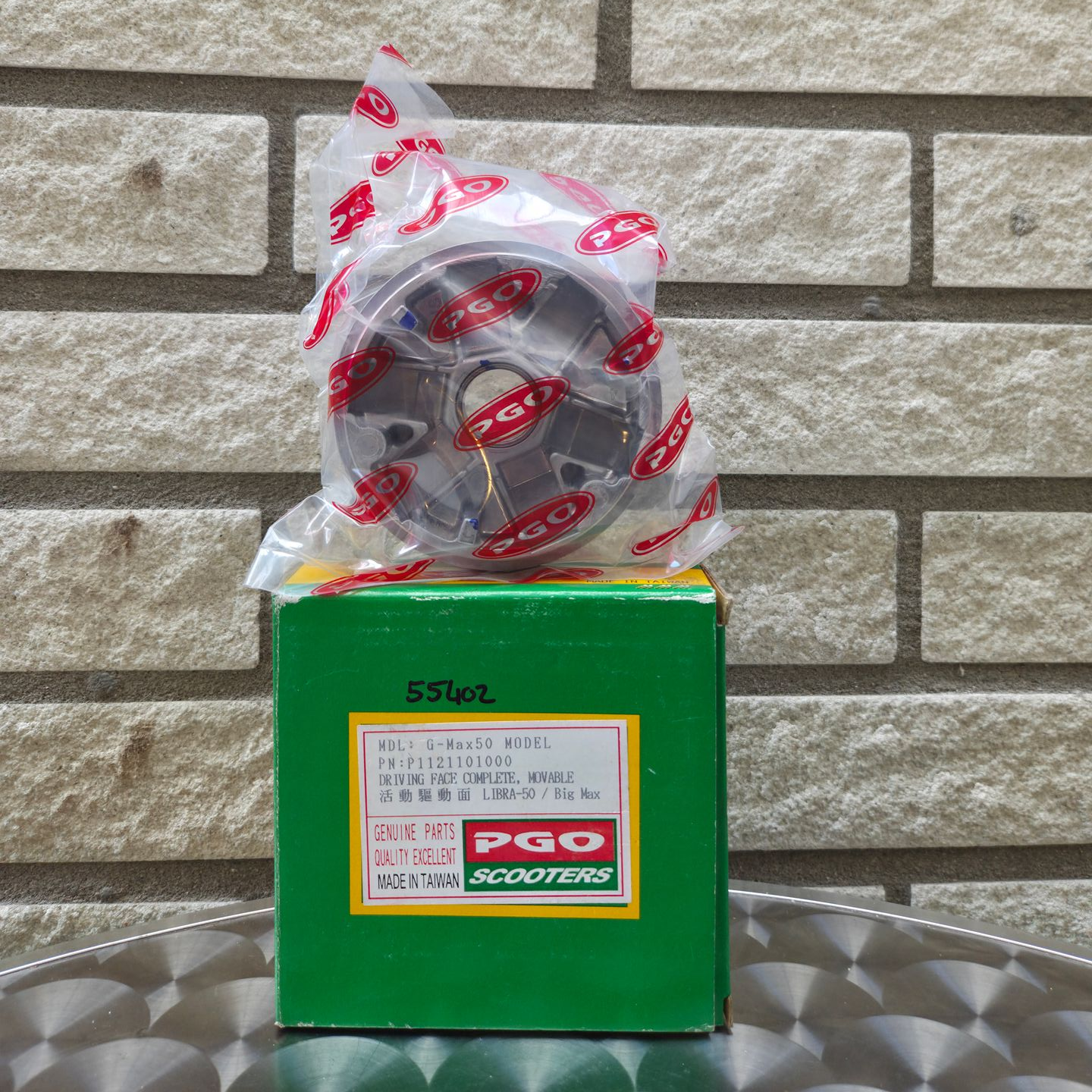 Een rood en zwart gemotoriseerd PGO-onderdeel, verpakt in plastic, staat op een groene doos tegen een bakstenen muur.