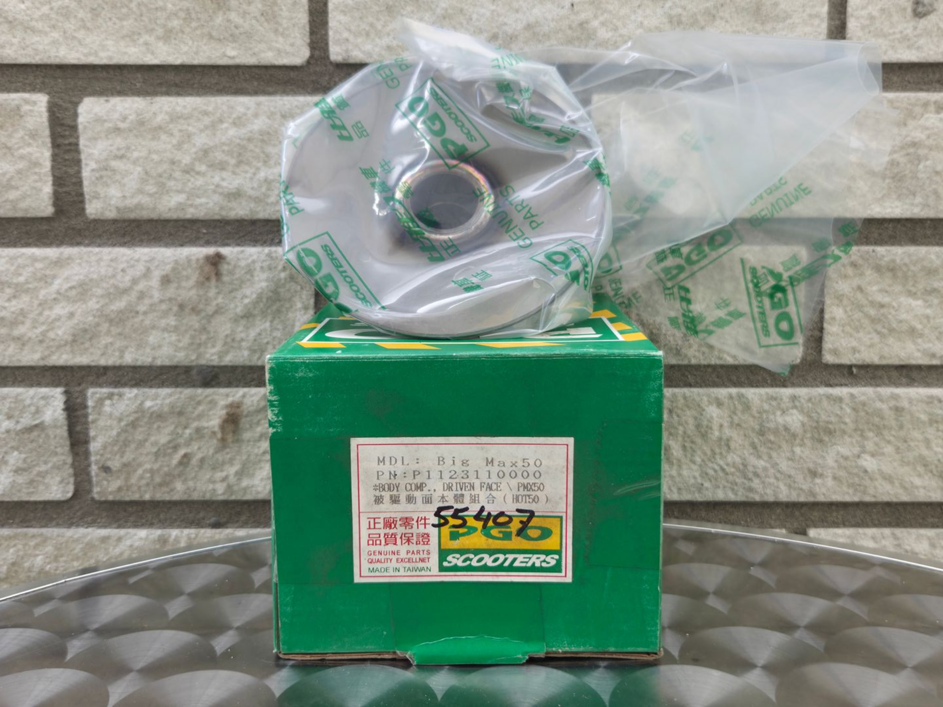 Groene doos met daarop een verzegelde plastic zak met onderdelen, tegen een achtergrond van een bakstenen muur.