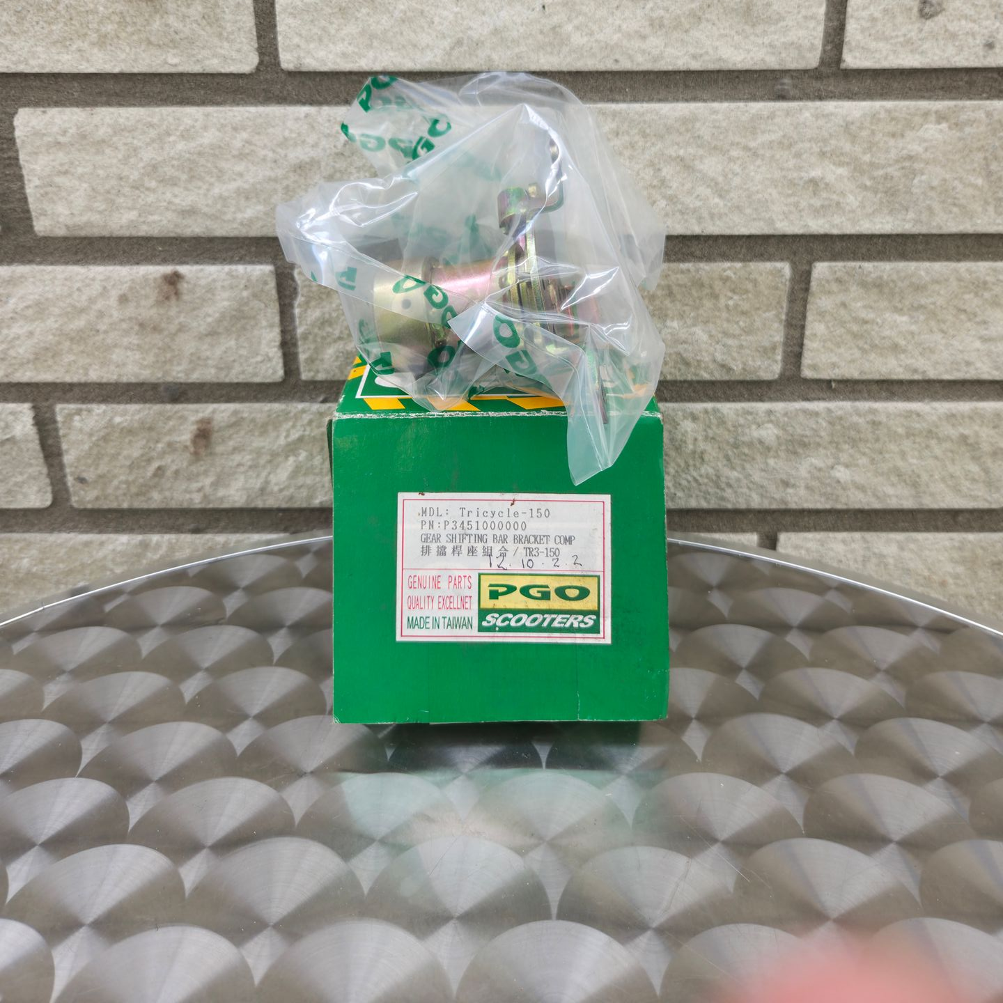 Een klein groen doosje met een in plastic verpakt speelgoedfiguurtje op een metalen tafel tegen een bakstenen muur.