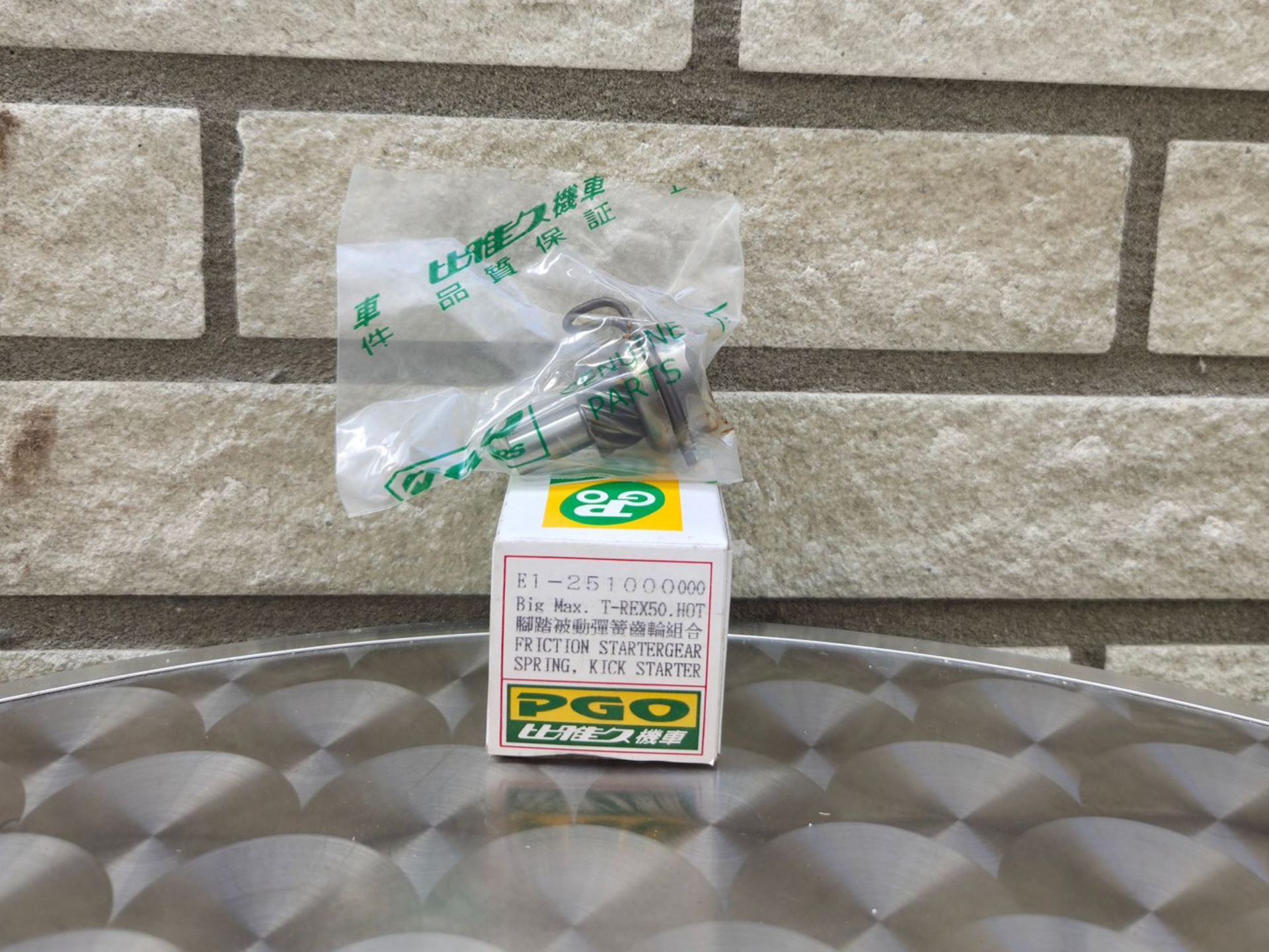 Klein, in een doosje verpakt product met een doorzichtig, groen dopje, omwikkeld met plastic, op een reflecterend metalen oppervlak tegen een bakstenen muur.