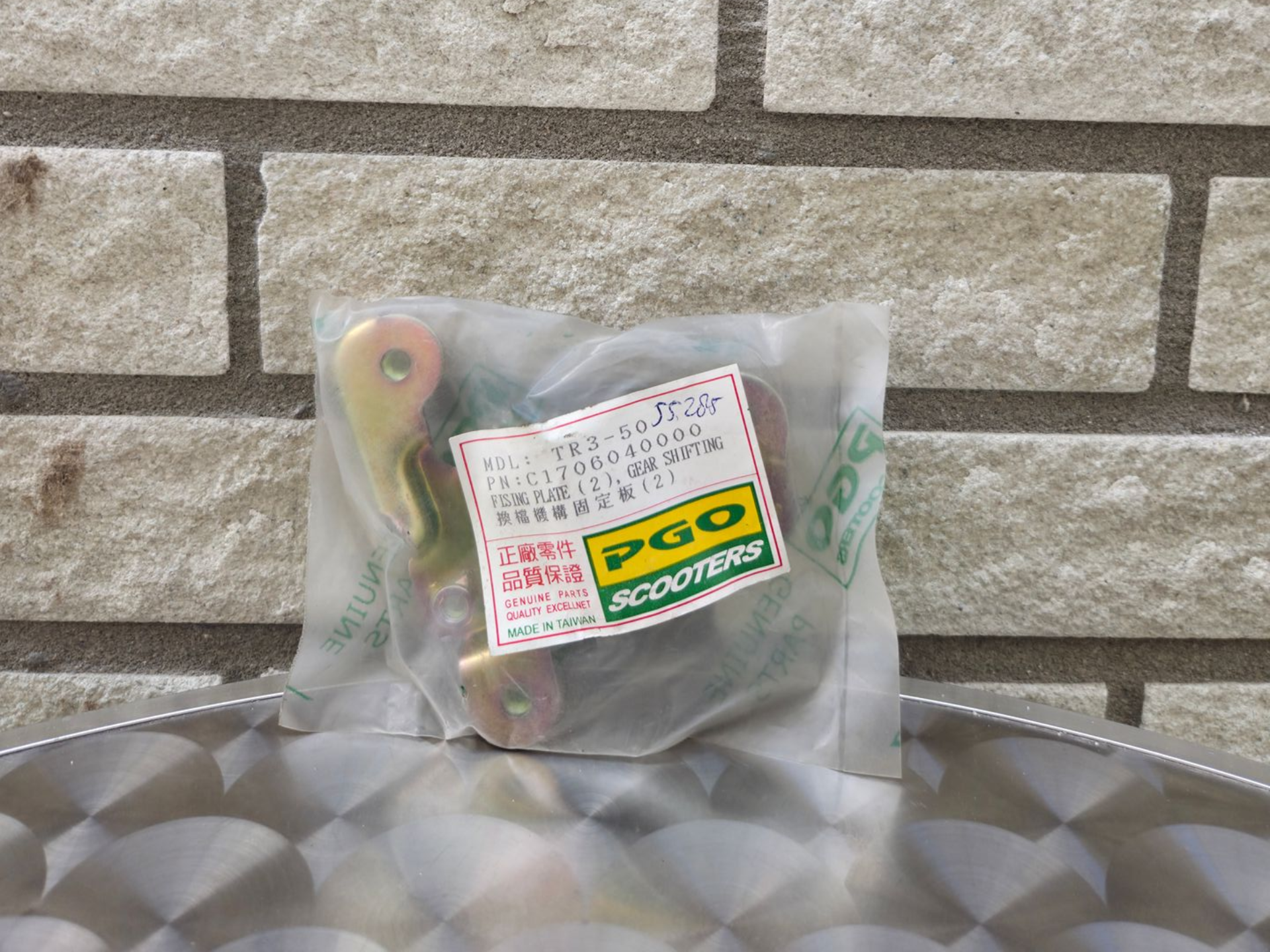 Een klein, doorzichtig plastic zakje met een etiket en een groen-geel label op een metalen oppervlak tegen een bakstenen muur.