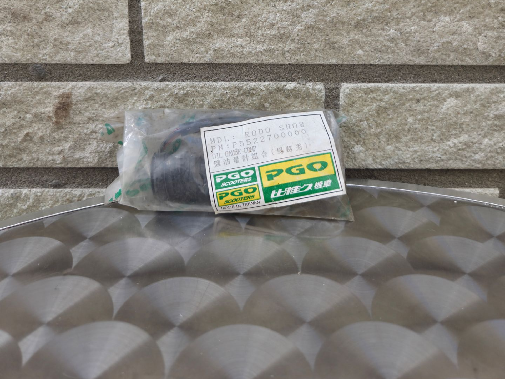 Een klein, groen-wit verpakt voorwerp dat op een metalen ondergrond met structuur tegen een bakstenen muur ligt.