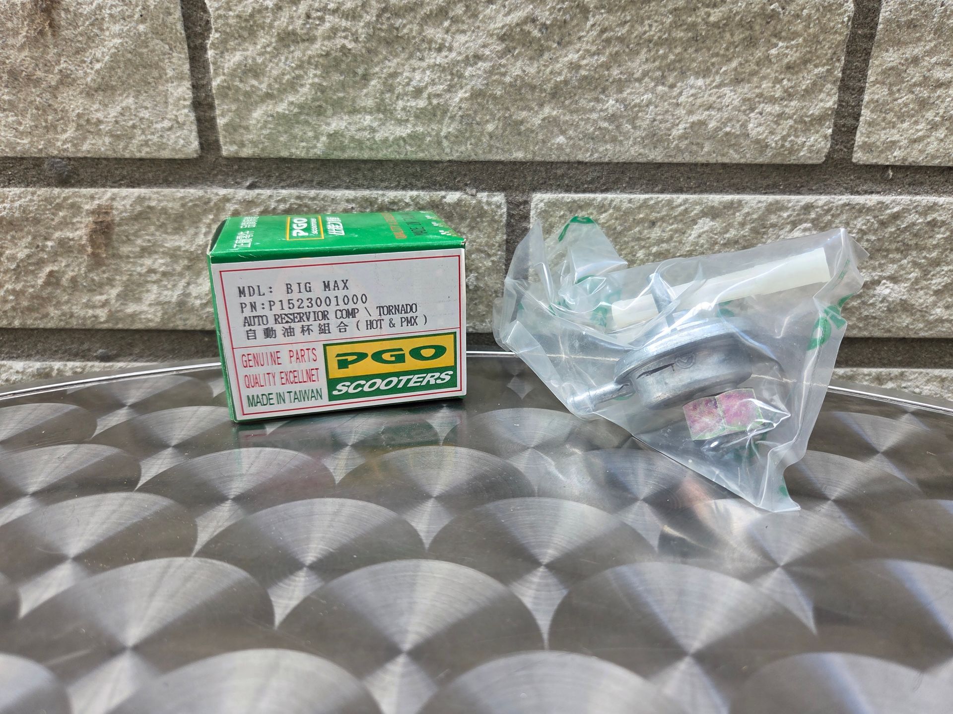 Een doos met een groen label en een doorzichtige plastic zak met kleine onderdelen op een metalen ondergrond met patroon, tegen een stenen muur.