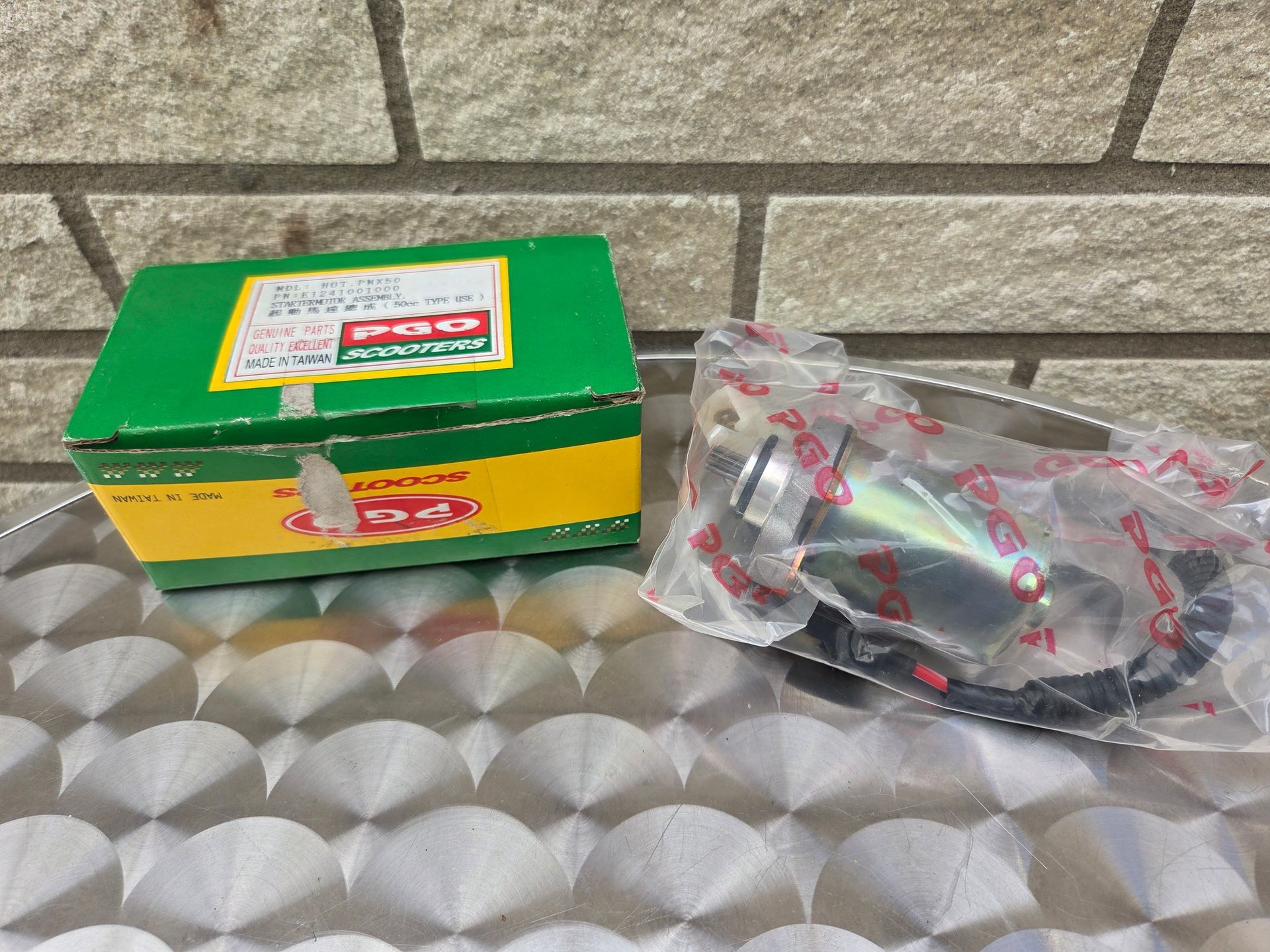 Een groen-gele doos naast een doorzichtige plastic zak met zwarte kabel op een metalen oppervlak met ruitpatroon.
