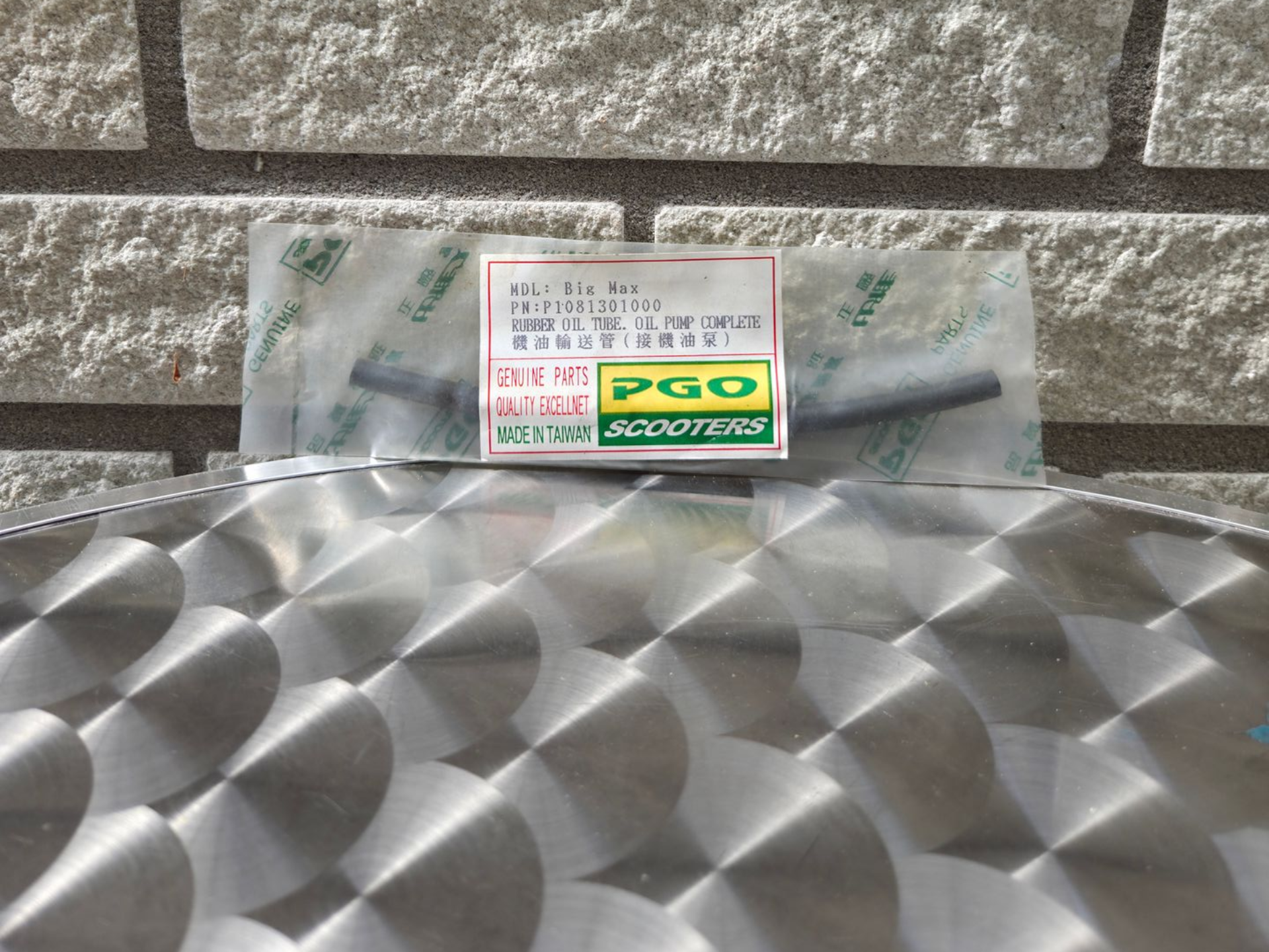 Klein, gelabeld pakketje met een geelgroene DGO-sticker, rustend op een metallic, patroonvormig oppervlak tegen een bakstenen muur.