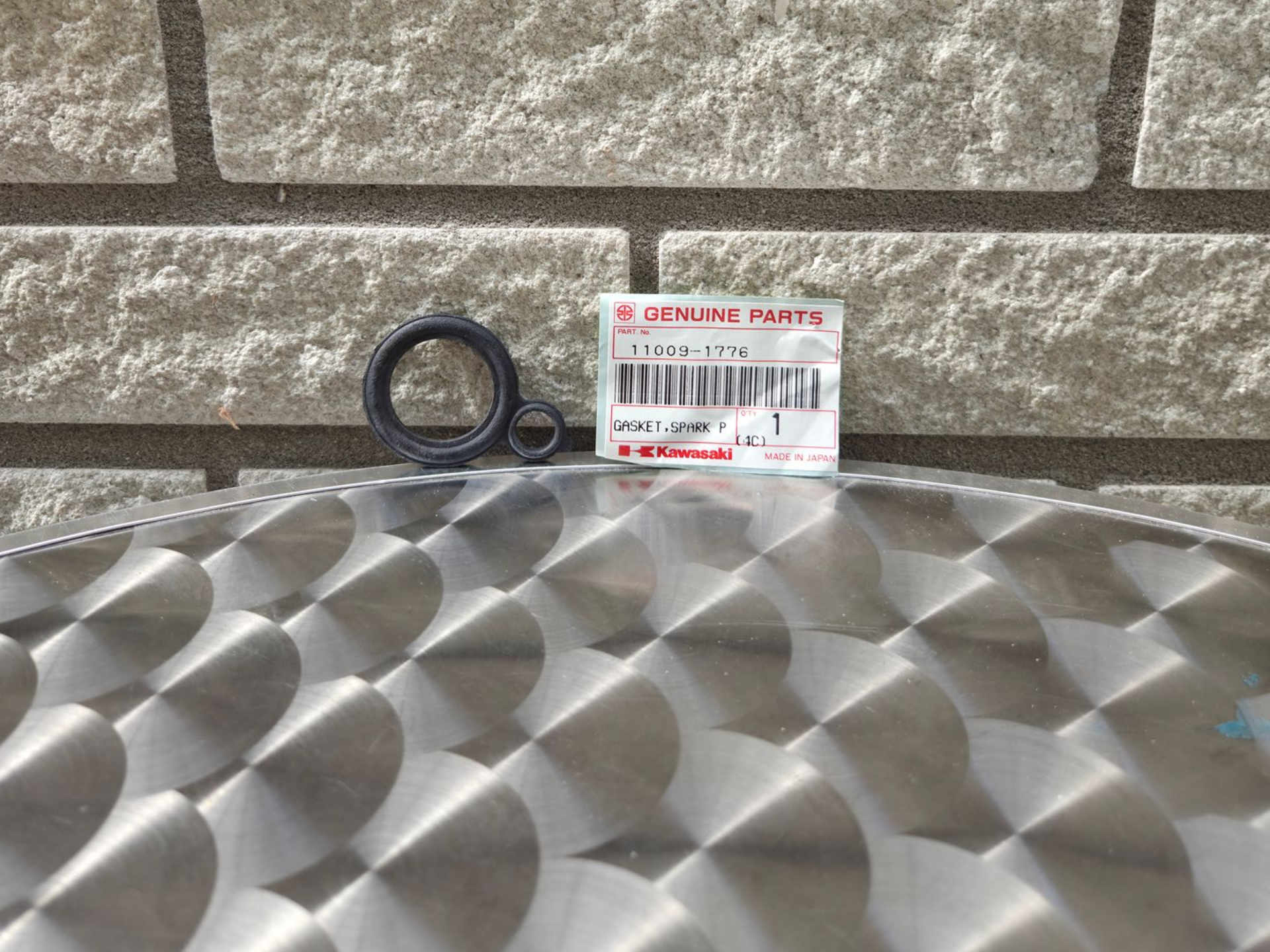 Zwarte ring en klein, gelabeld pakketje op een zilverkleurige, diamantvormige ondergrond tegen een grijze stenen muur.