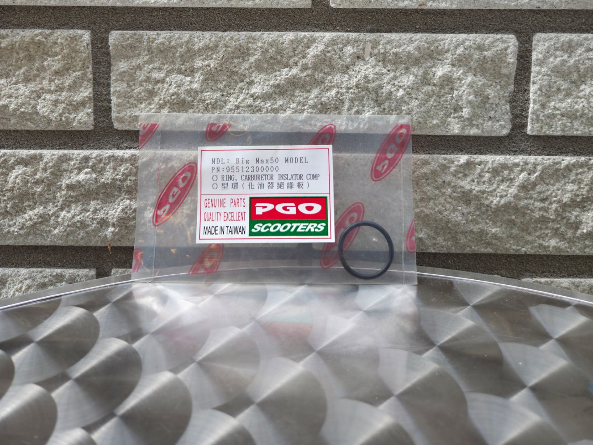 Doorzichtige plastic zak met rood-witte PGO-sticker en zwarte ring, rustend op een grijs, geruit oppervlak tegen een stenen muur.