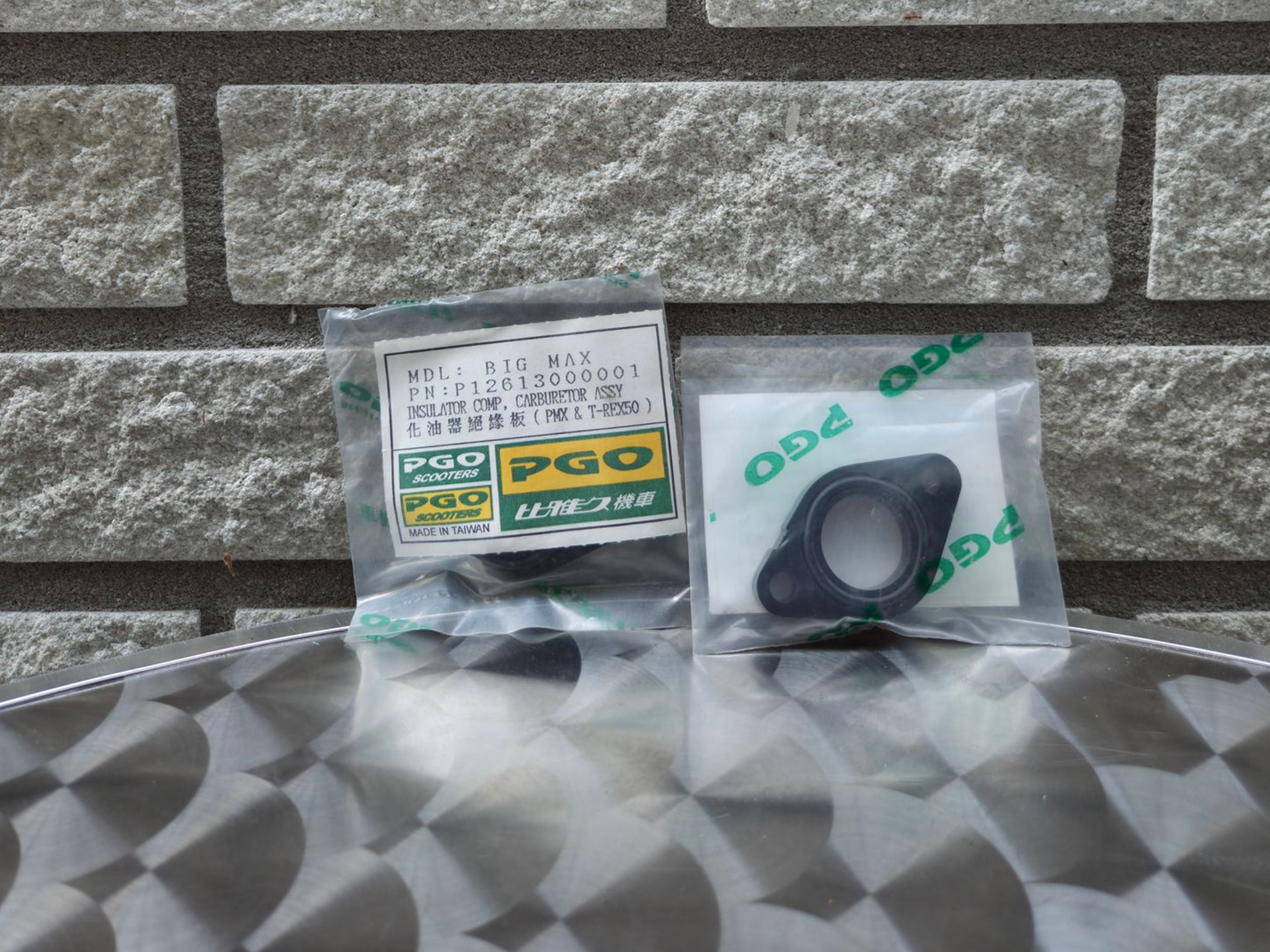 Twee verzegelde PGO-onderdelenpakketten op een metalen oppervlak voor een stenen muur.