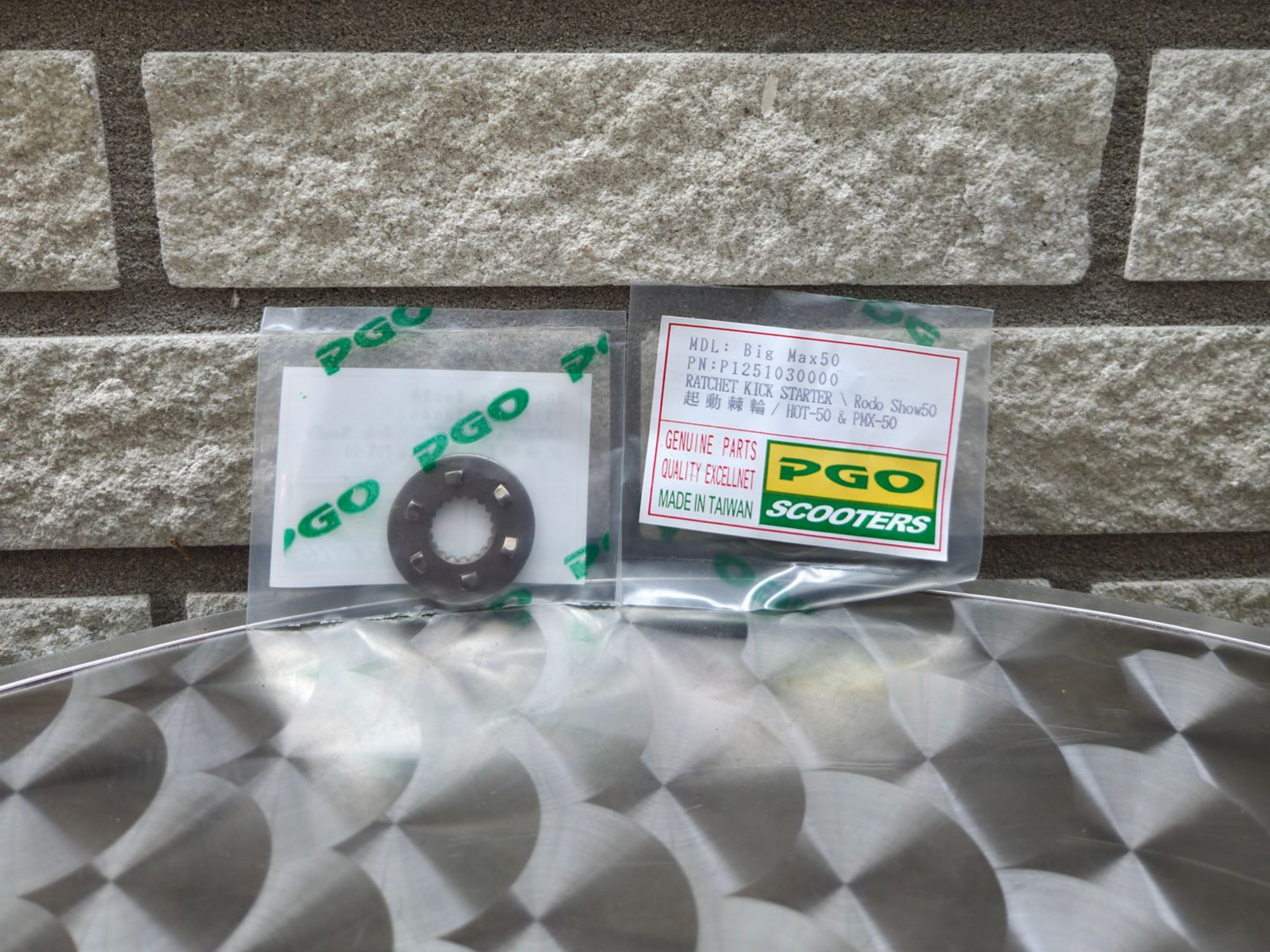 Twee verpakte auto-onderdelen op een metalen oppervlak met patroon tegen een stenen muur.