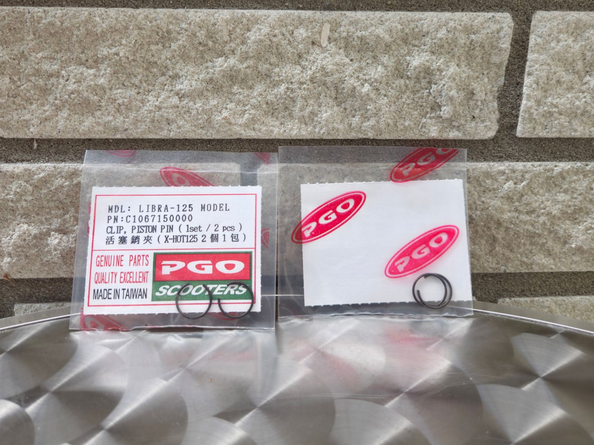Twee kleine doosjes met het PGO-logo op een zilverkleurige, gestructureerde ondergrond tegen een stenen muur.