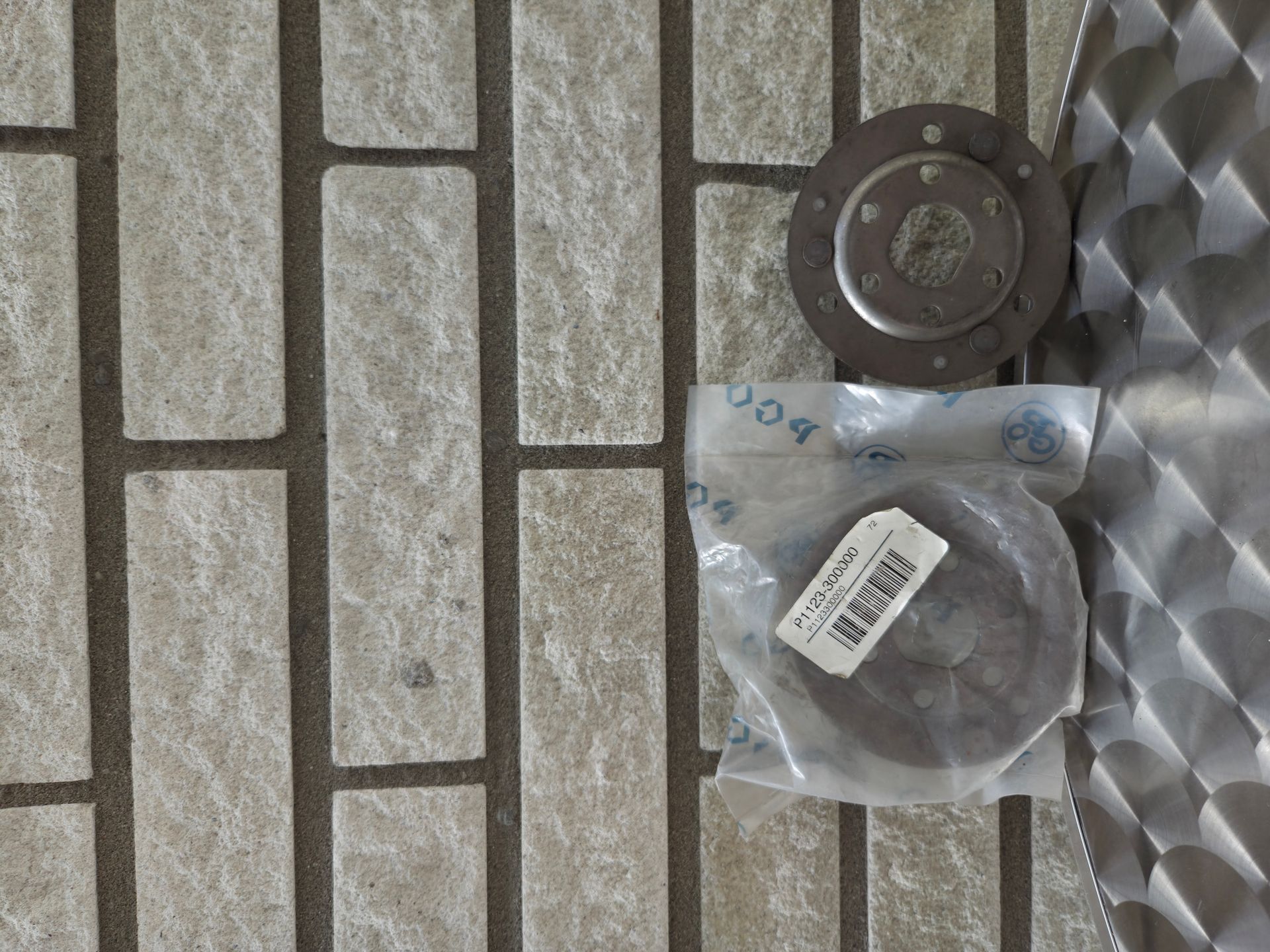 Twee ronde metalen onderdelen in doorzichtige plastic zakken naast een grijze bakstenen muur en een tegelvloer met patroon.