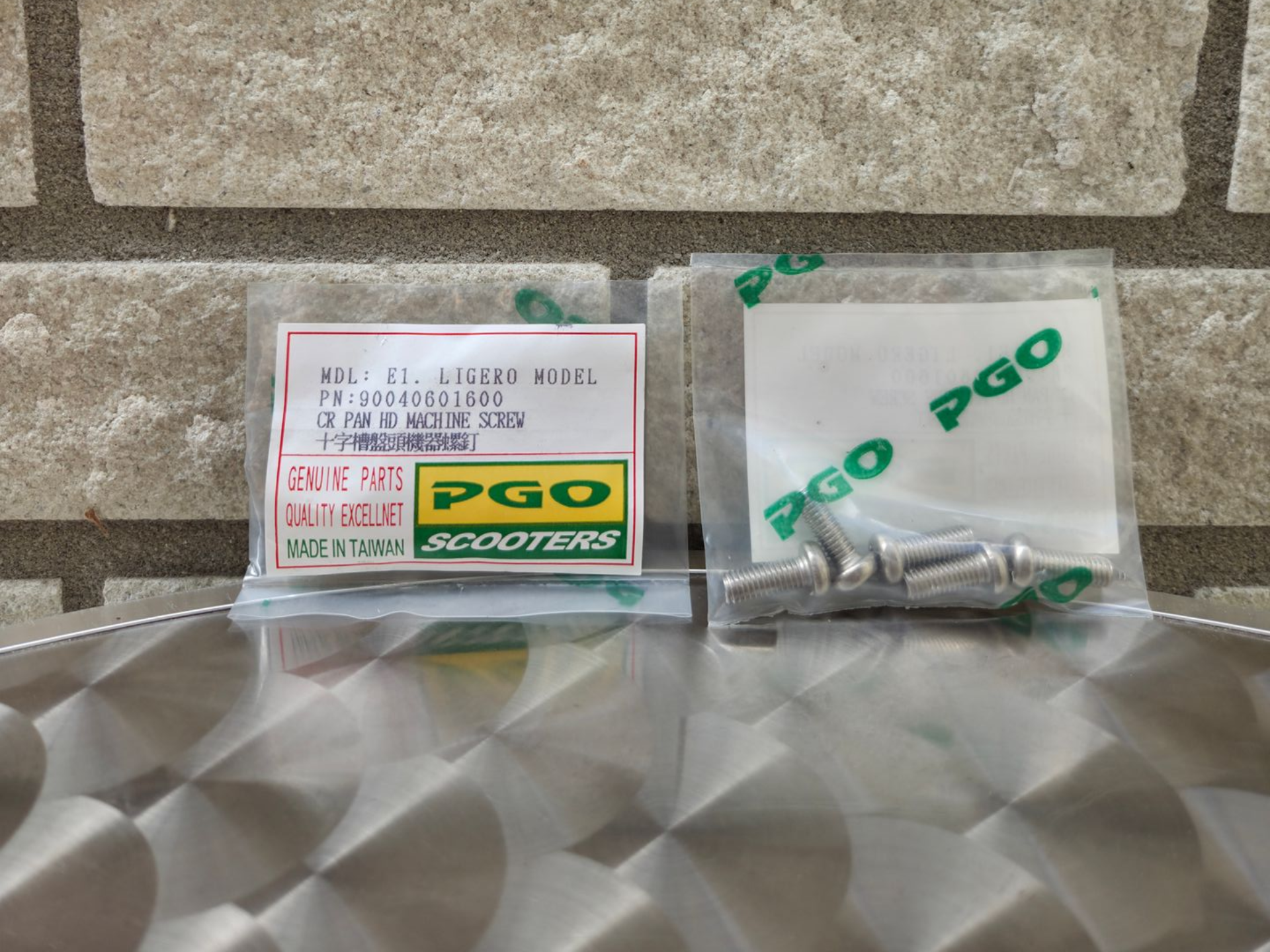 Een tas met het PGO-logo en kleine metalen onderdelen, naast een doos met onderdelen en een label, op een tafelblad met textuur.