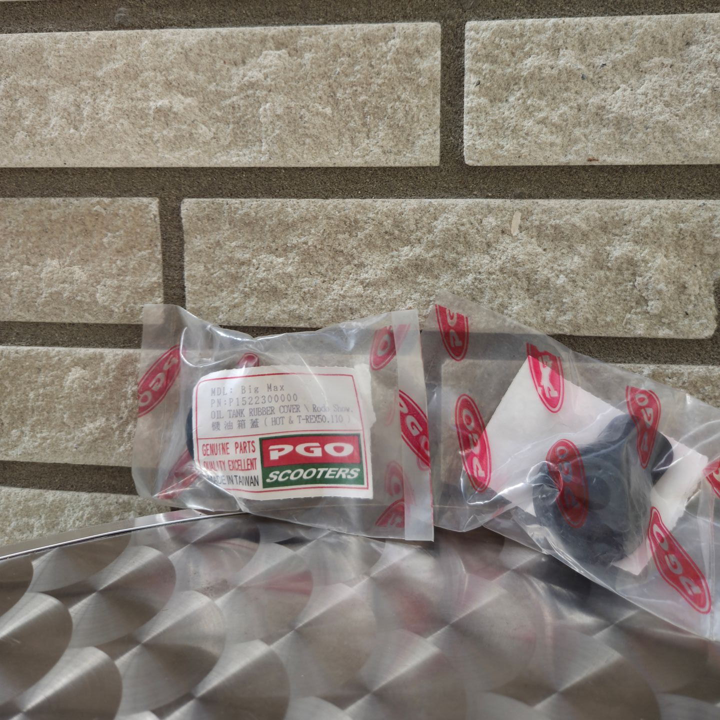 Twee kleine onderdelen in doorzichtige plastic zakjes op een zilverkleurig, diamantvormig oppervlak tegen een stenen muur.