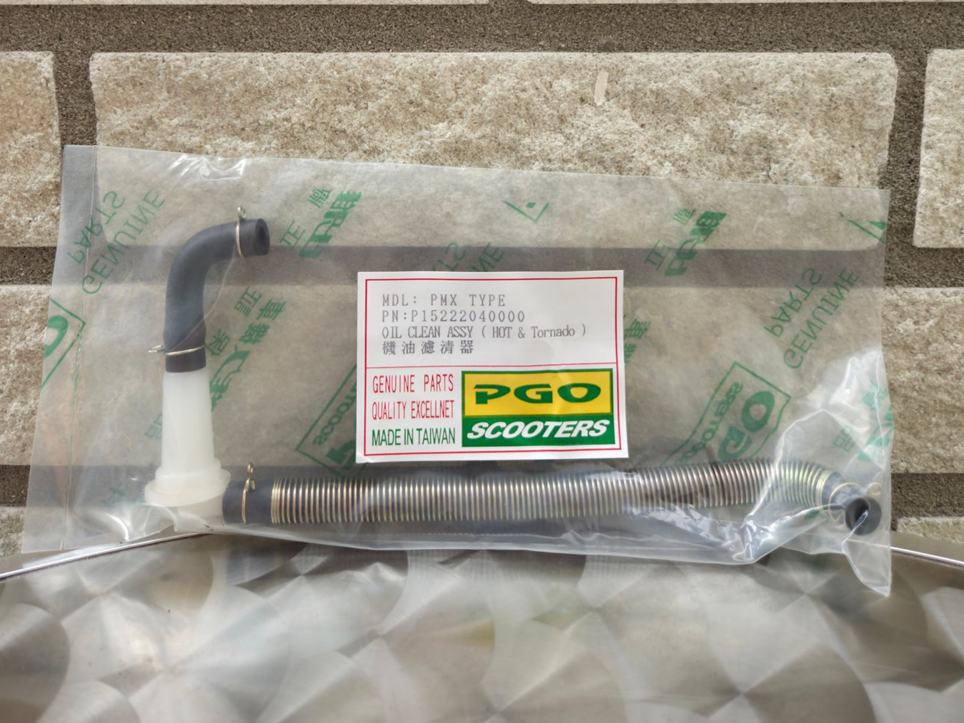 Een plastic scooteronderdeel in een doorzichtige zak met een PGO Scooters-label op een gestructureerd oppervlak.