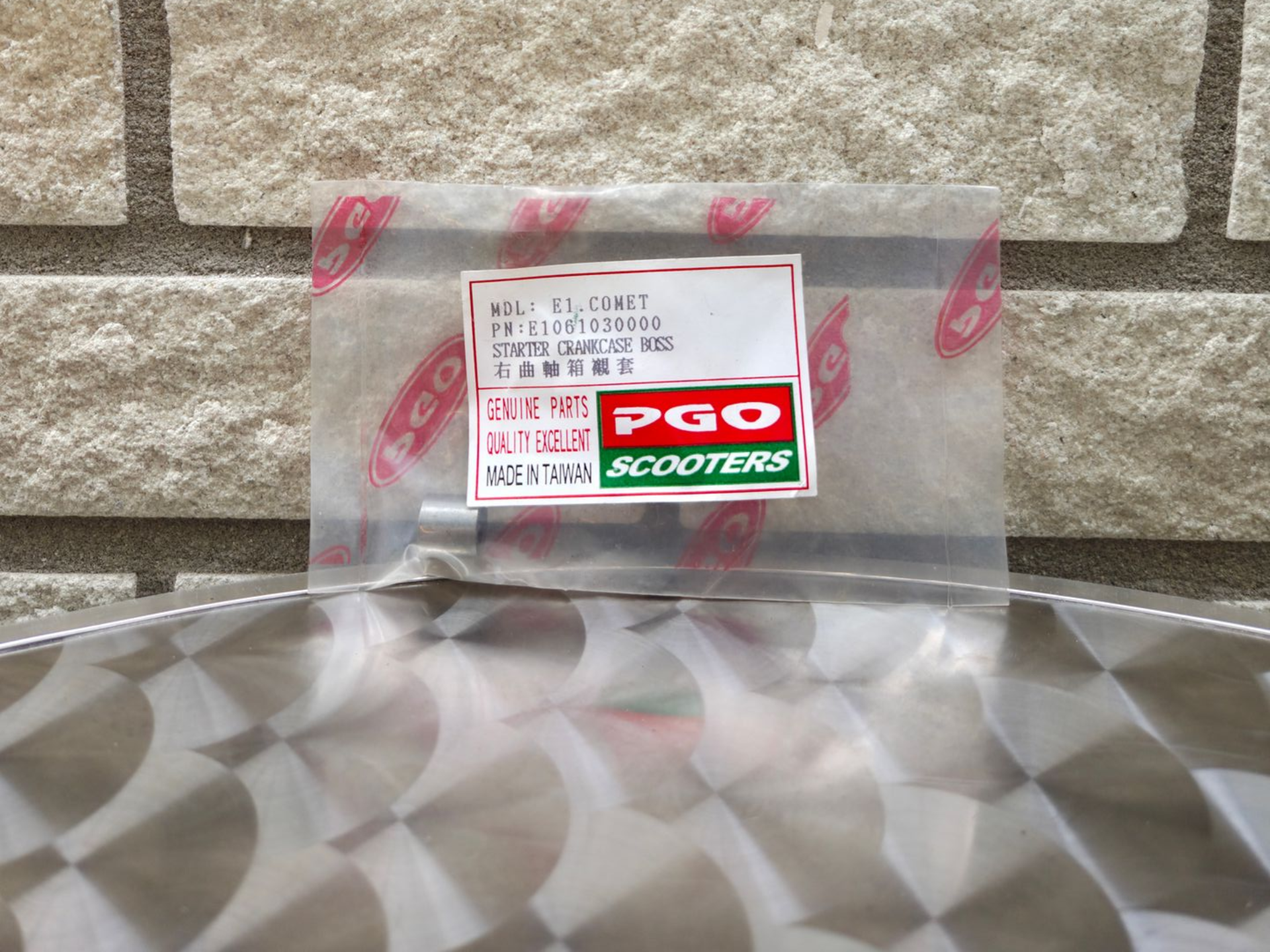 Een doorzichtige plastic zak met een PGO Scooters-label op een tafelblad met patroon, tegen een bakstenen muur.