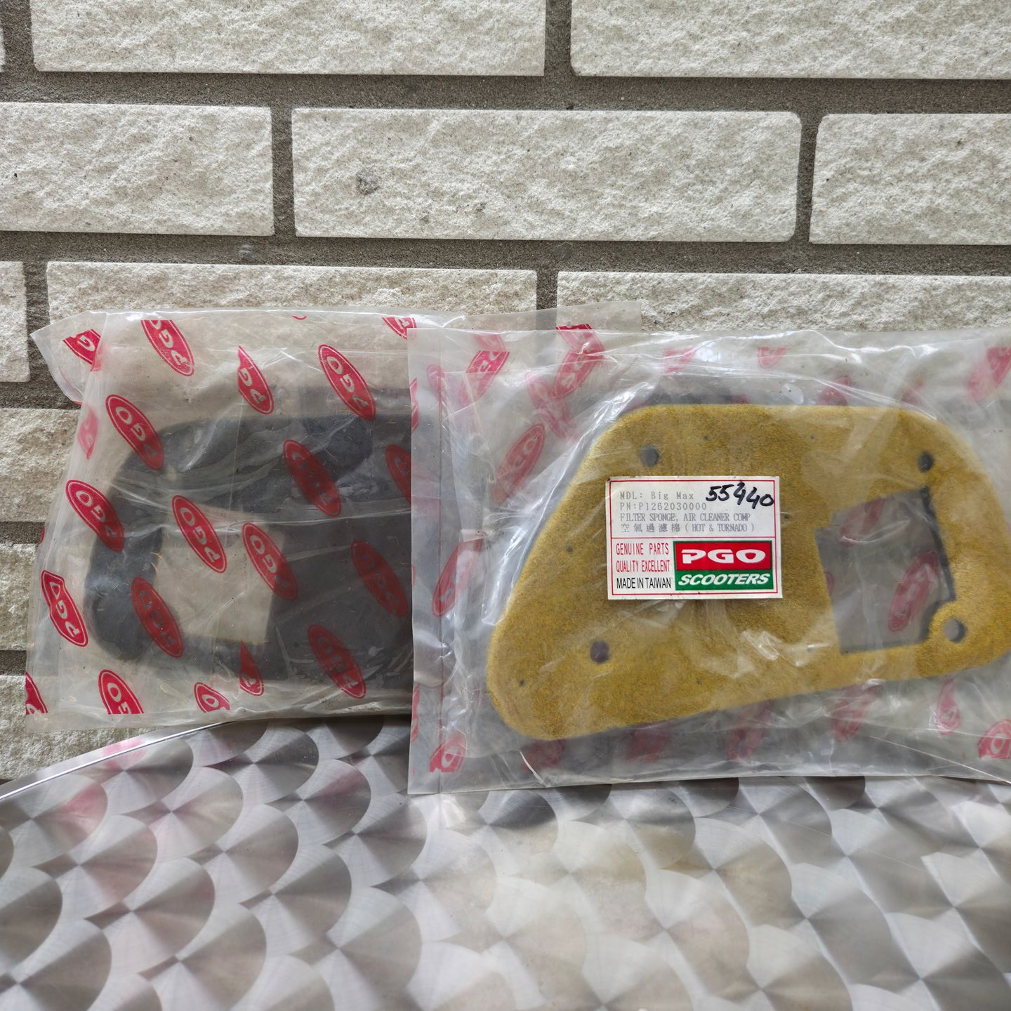 Twee verpakte gele driehoekige onderdelen en een donkerrood component op een oppervlak met patroon tegen een bakstenen muur.