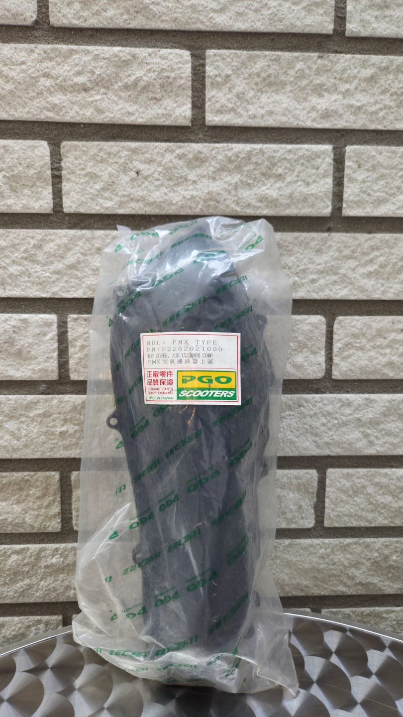 Een zwart voorwerp, verpakt in een doorzichtige plastic verpakking, staat rechtop tegen een bakstenen muur.