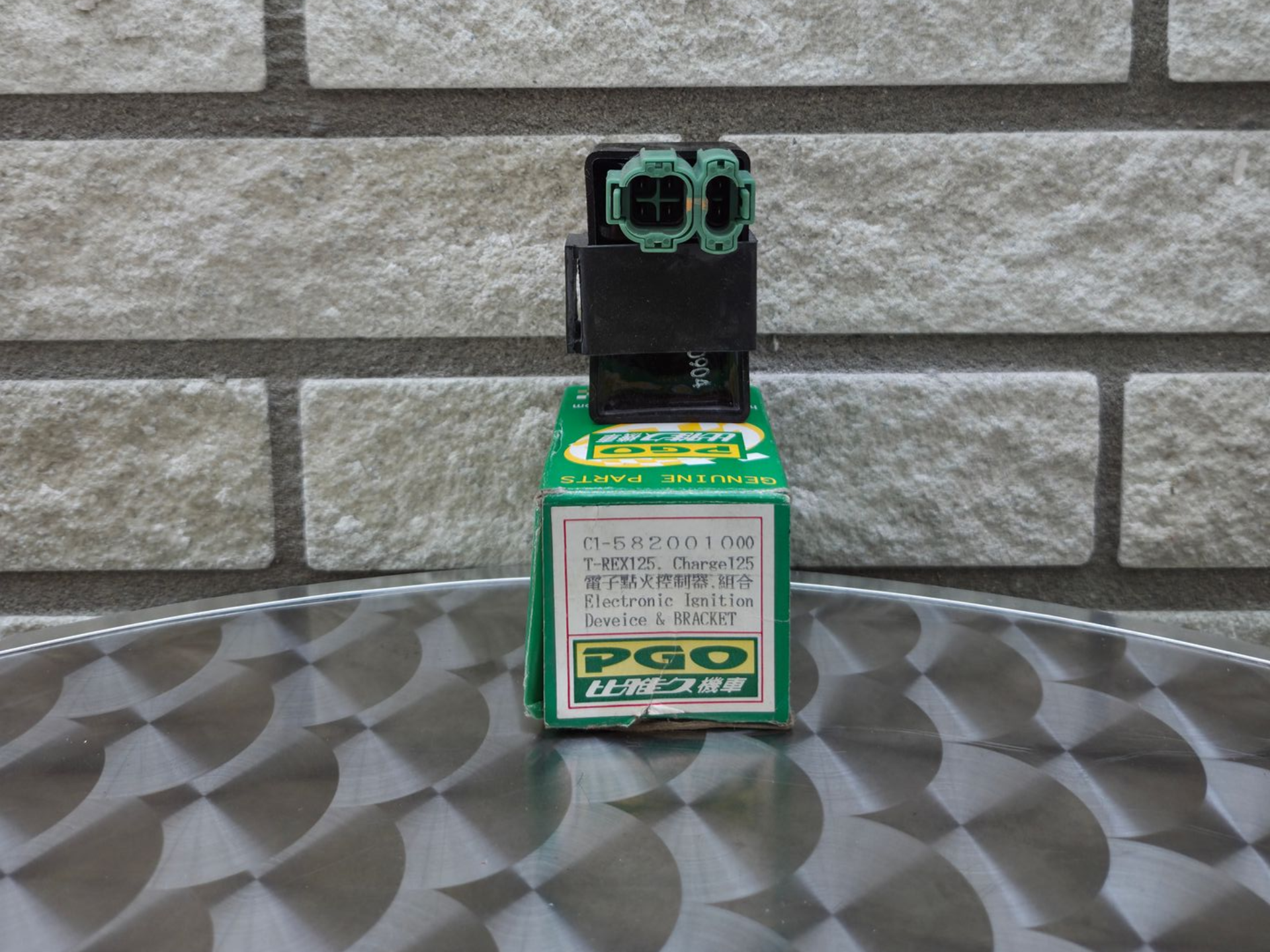 Een klein groen-zwart speelgoedrobotje staat op een groene doos tegen een grijze bakstenen muur.