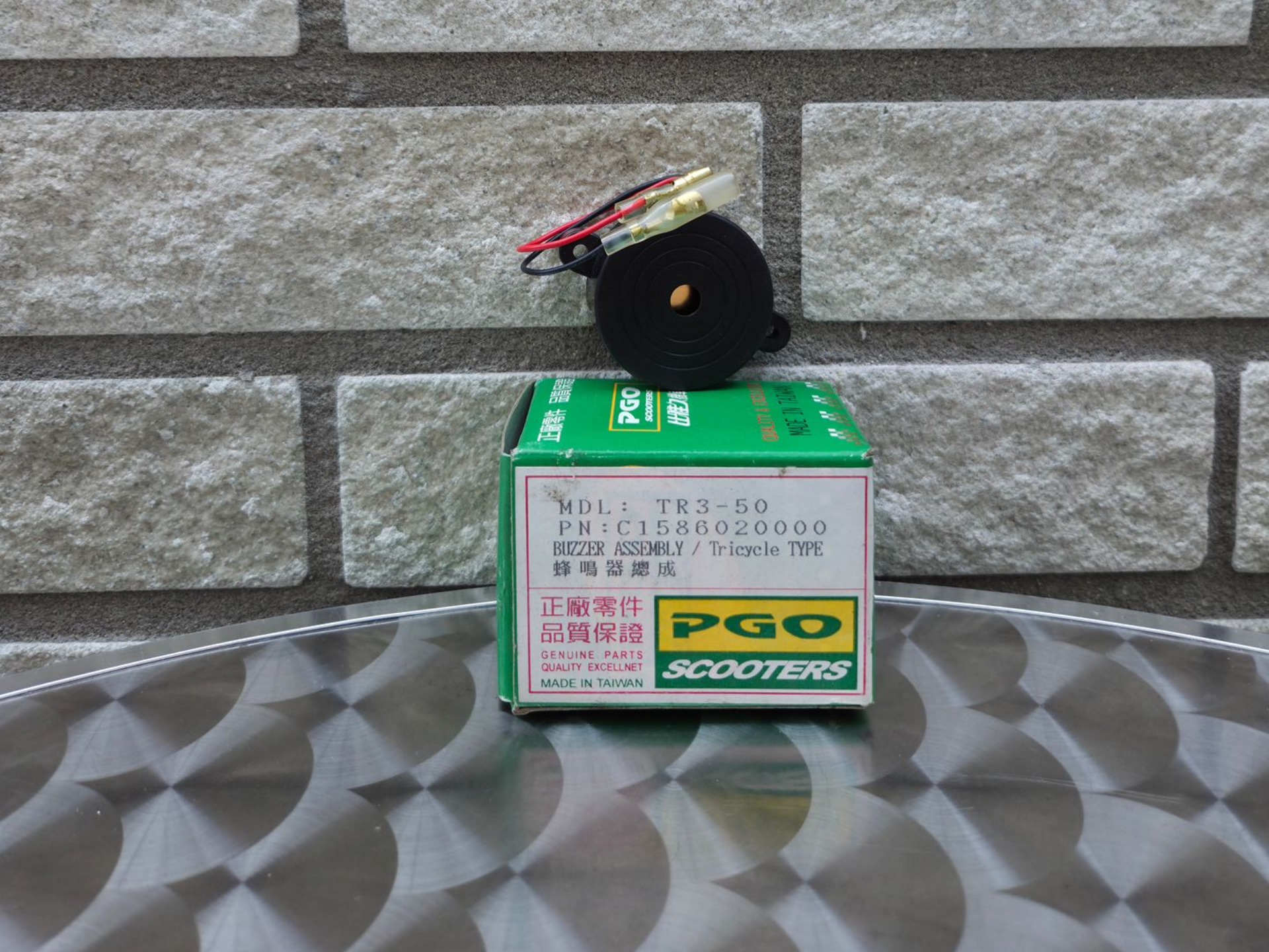 Een kleine zwart-rode speelgoedstep op een groene doos, die op een metalen ondergrond met patroon staat tegen een bakstenen muur.