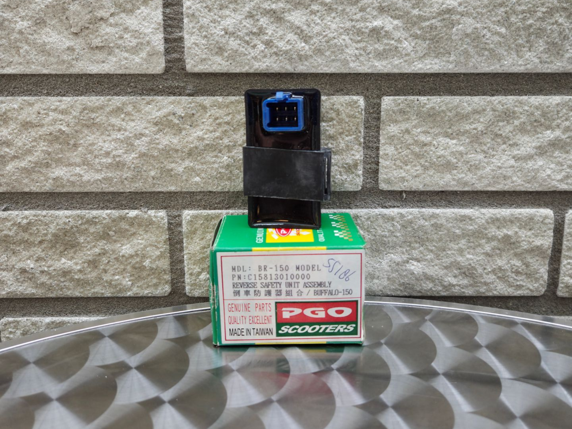 Een klein groen doosje met het opschrift PGO en een zwart/blauwe sensor erop, tegen een bakstenen muur.