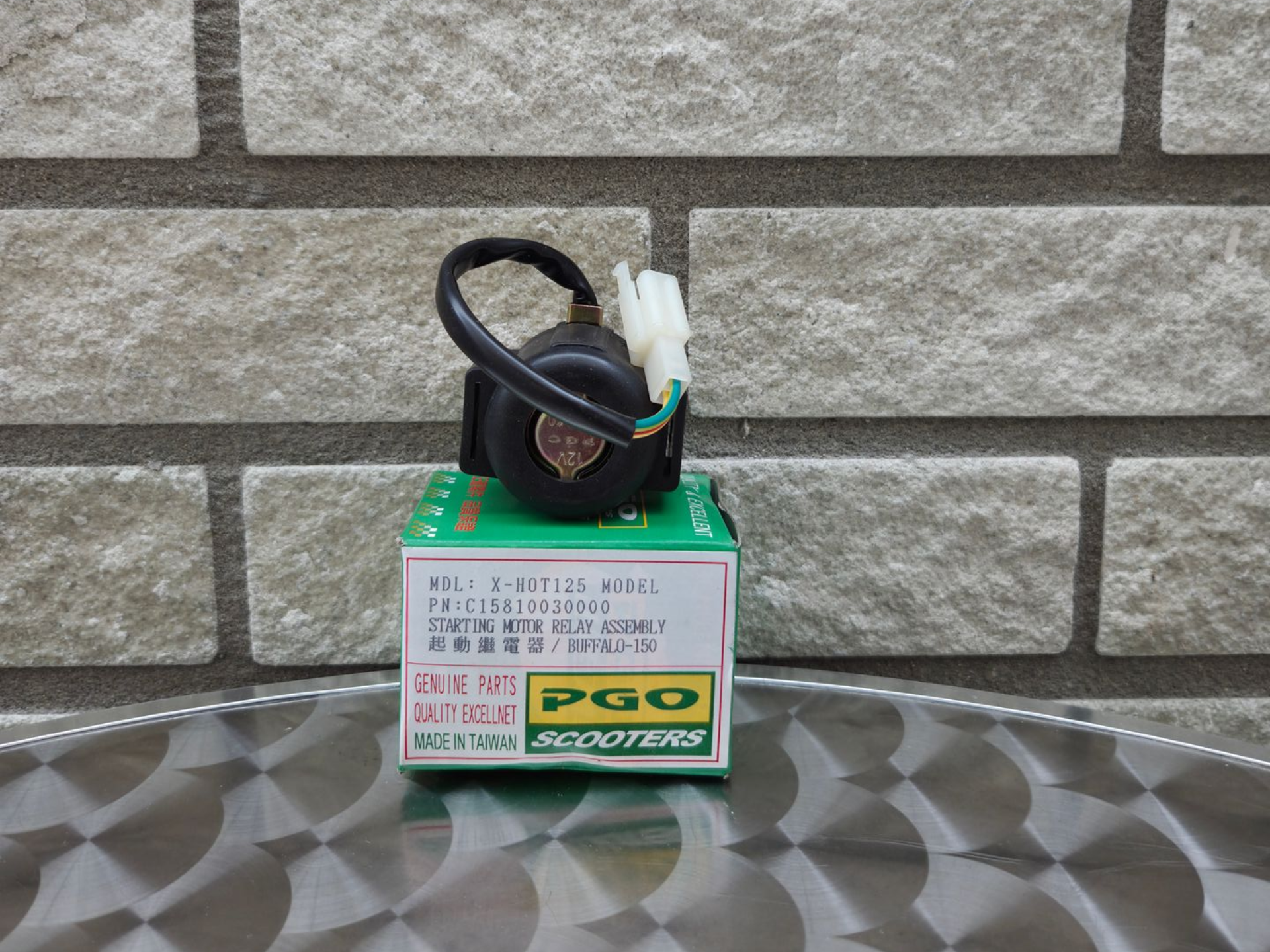 Een opgerolde zwarte kabel met witte connector ligt op een groene PCO-apparaatdoos tegen een bakstenen muur.