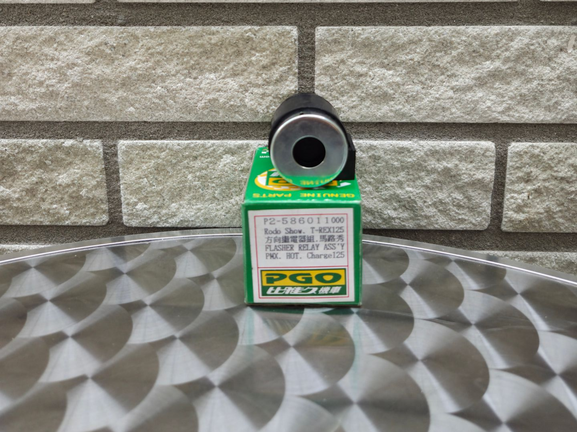 Een klein groen-wit doosje met daarop een zwart, cirkelvormig, camera-achtig object, op een metalen ondergrond tegen een bakstenen muur.