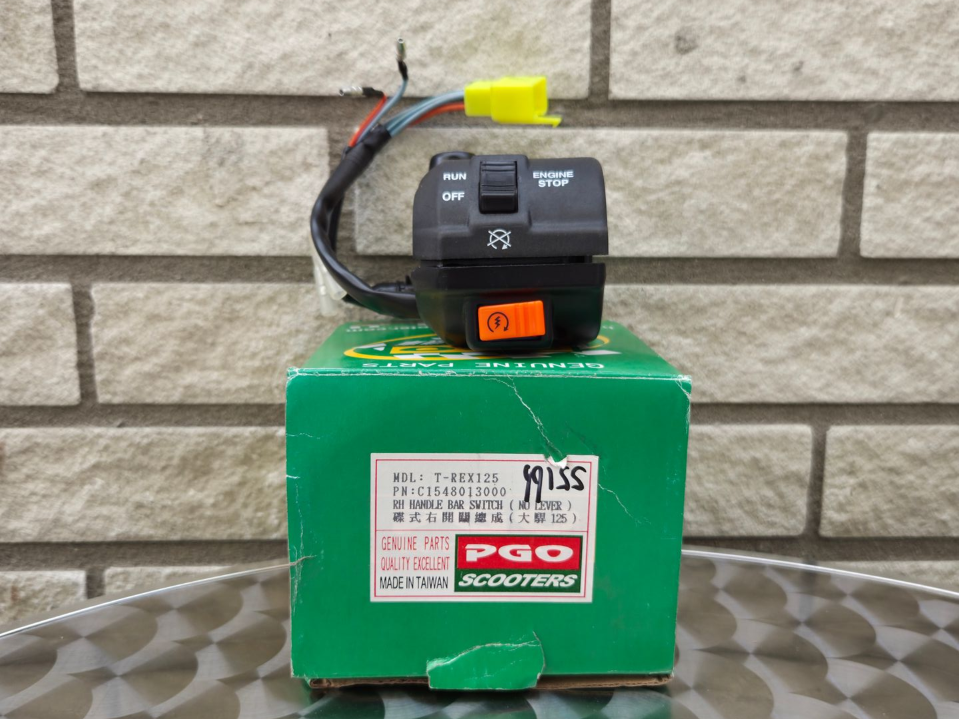 Zwarte schakelaar gemonteerd op een groene doos tegen een bakstenen muur, met bedrading en een gele connector erbovenop.