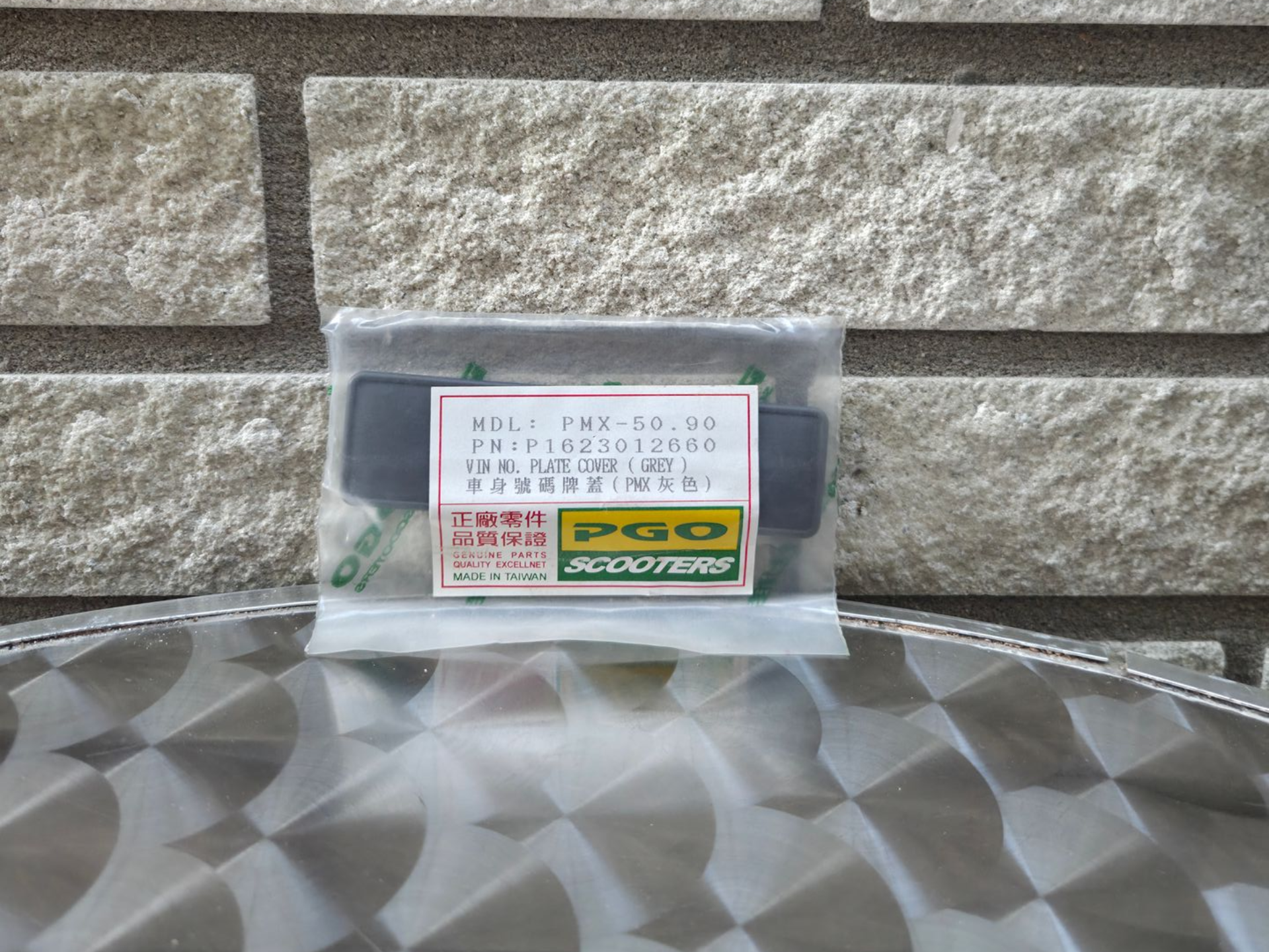 Klein, doorzichtig plastic doosje met een wit etiket en een geelgroene sticker, dat buiten op een tafel met een patroon staat.
