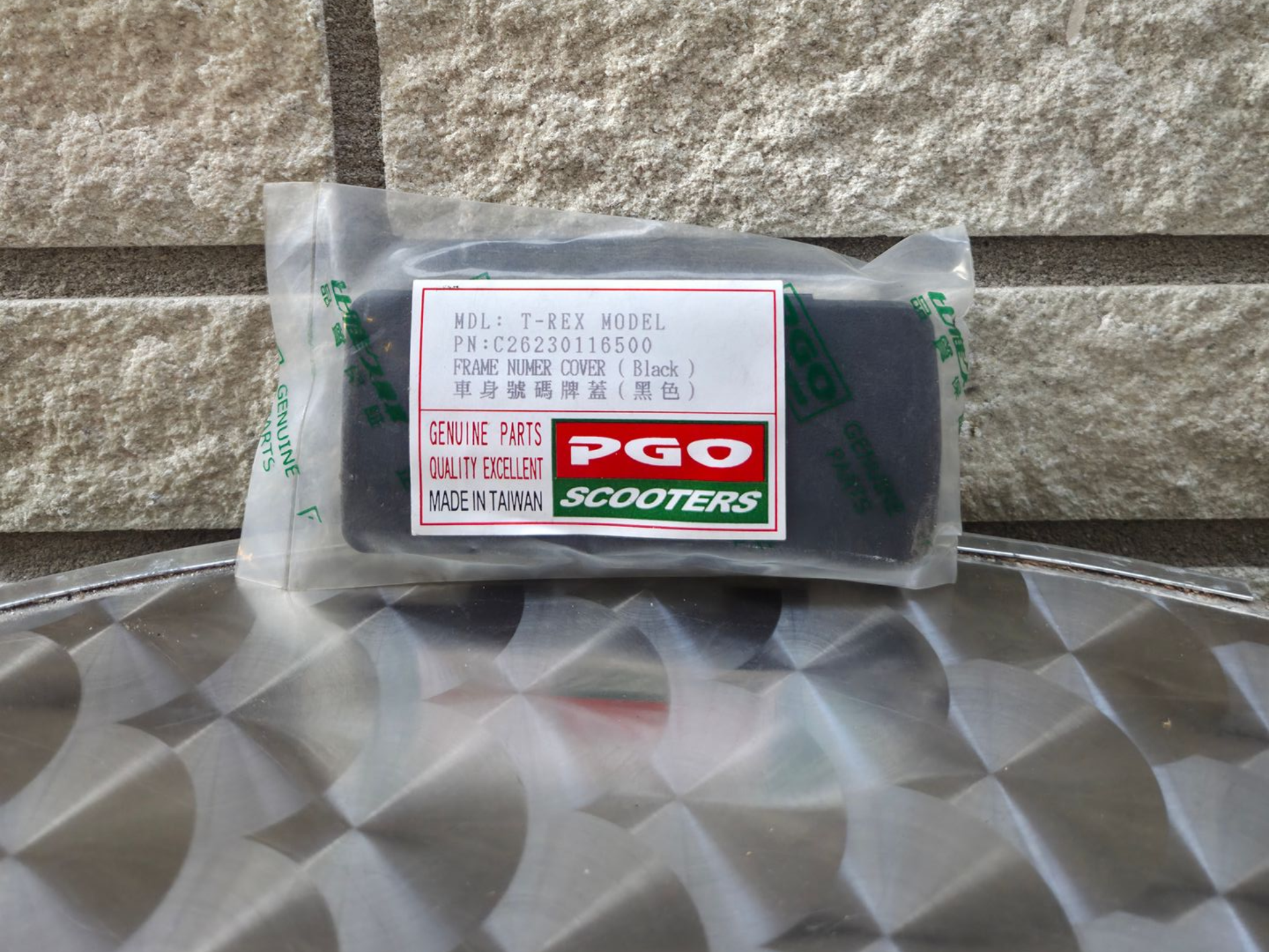 Doorzichtige plastic zak met een rood en groen PGO Scooters-label op een grijs, bedrukt oppervlak.