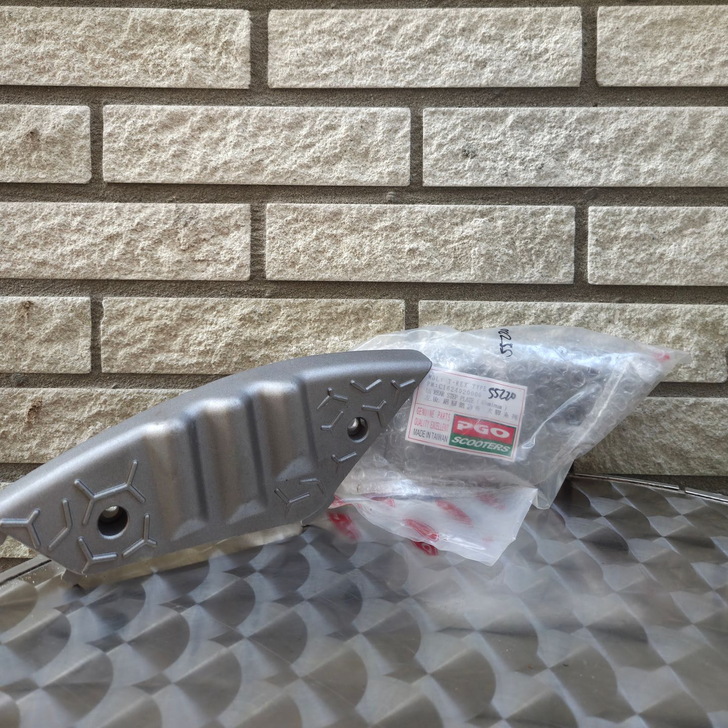 Zilverkleurig metalen onderdeel met een doorzichtig plastic zakje met kleine onderdelen op een ondergrond met patroon, tegen een achtergrond van een bakstenen muur.