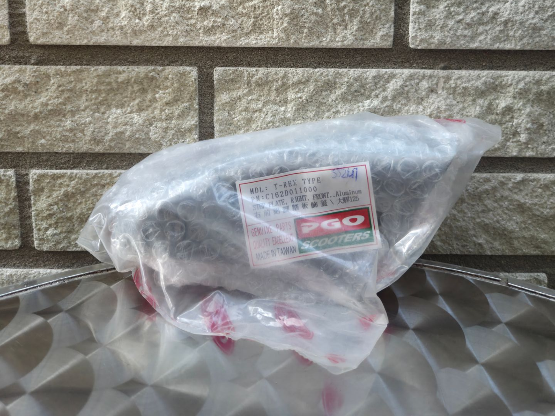 Een in plastic verpakt voorwerp met een rood etiket, rustend op een metalen tafel tegen een bakstenen muur.