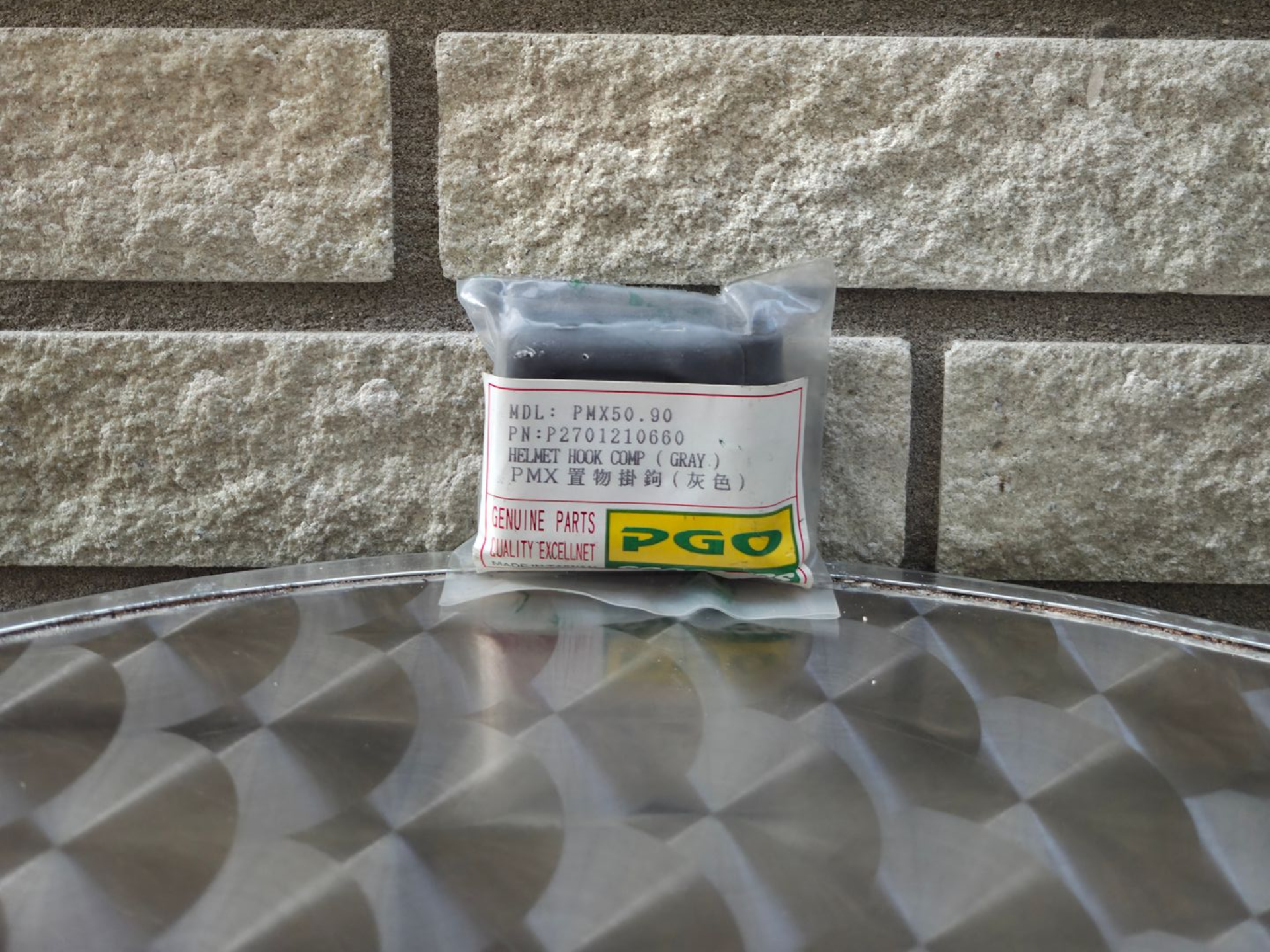 Een klein, verpakt artikel met het PGO-logo, geplaatst op een reflecterend metalen oppervlak tegen een stenen muur.
