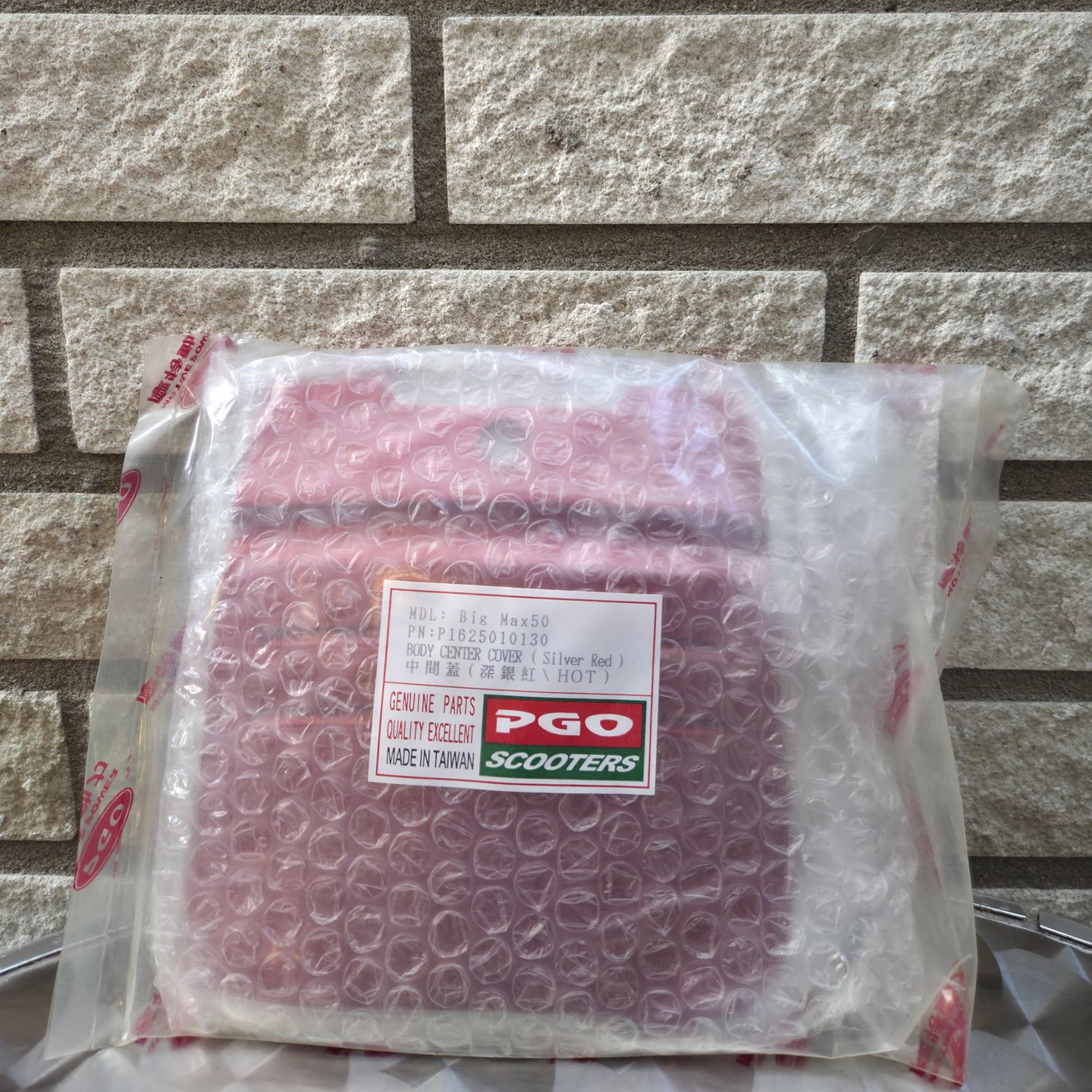 Een roze scooteronderdeel, verpakt in bubbeltjesplastic en vervolgens in een doorzichtige plastic zak, staat tegen een bakstenen muur.