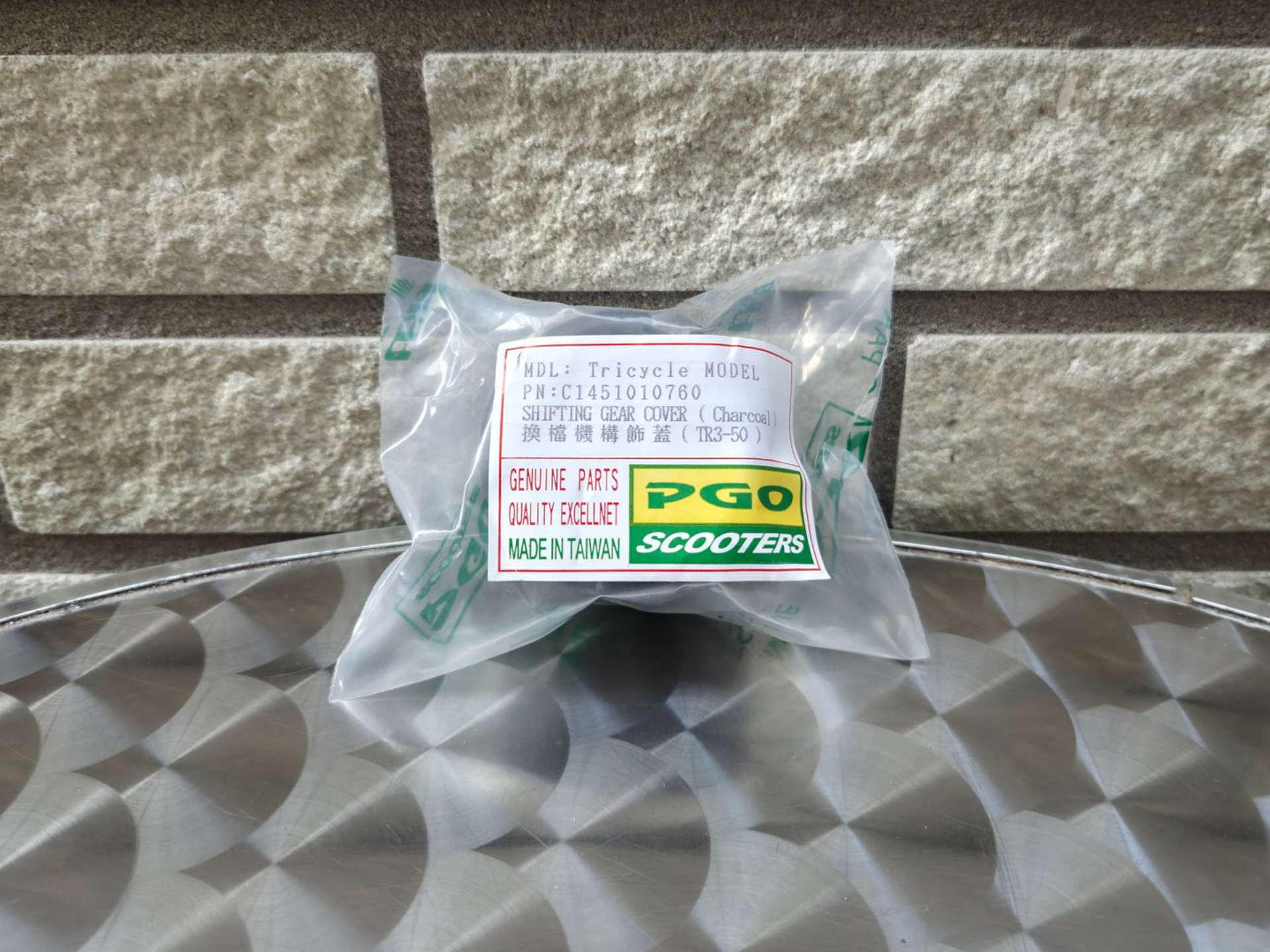 Een klein, doorzichtig plastic zakje met PCG-scooteronderdelen op een geruite tafel tegen een stenen muur.