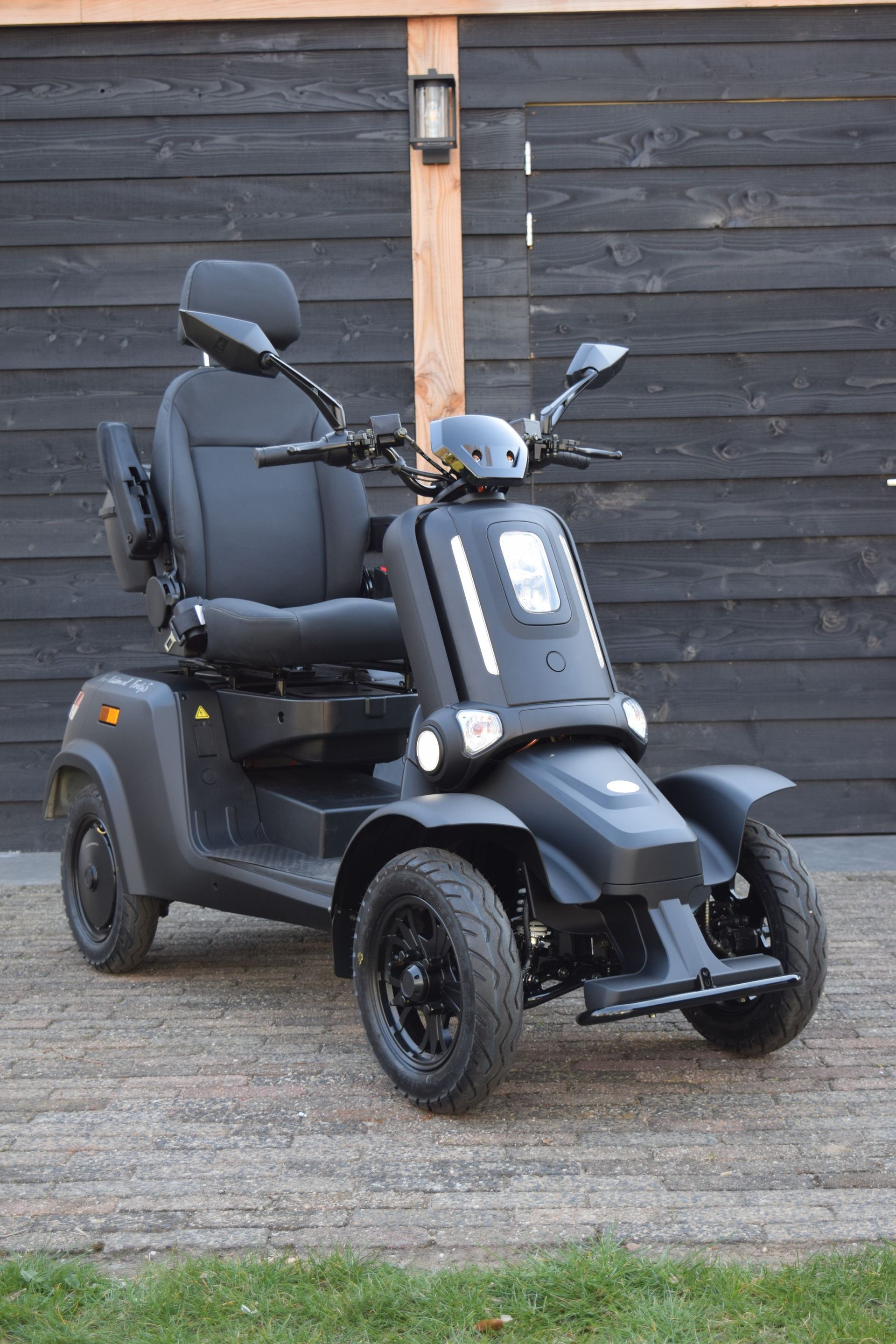 Zwarte scootmobiel buiten geparkeerd. Donkergrijze lak, voorzien van koplampen, comfortabele zitting en spiegels.