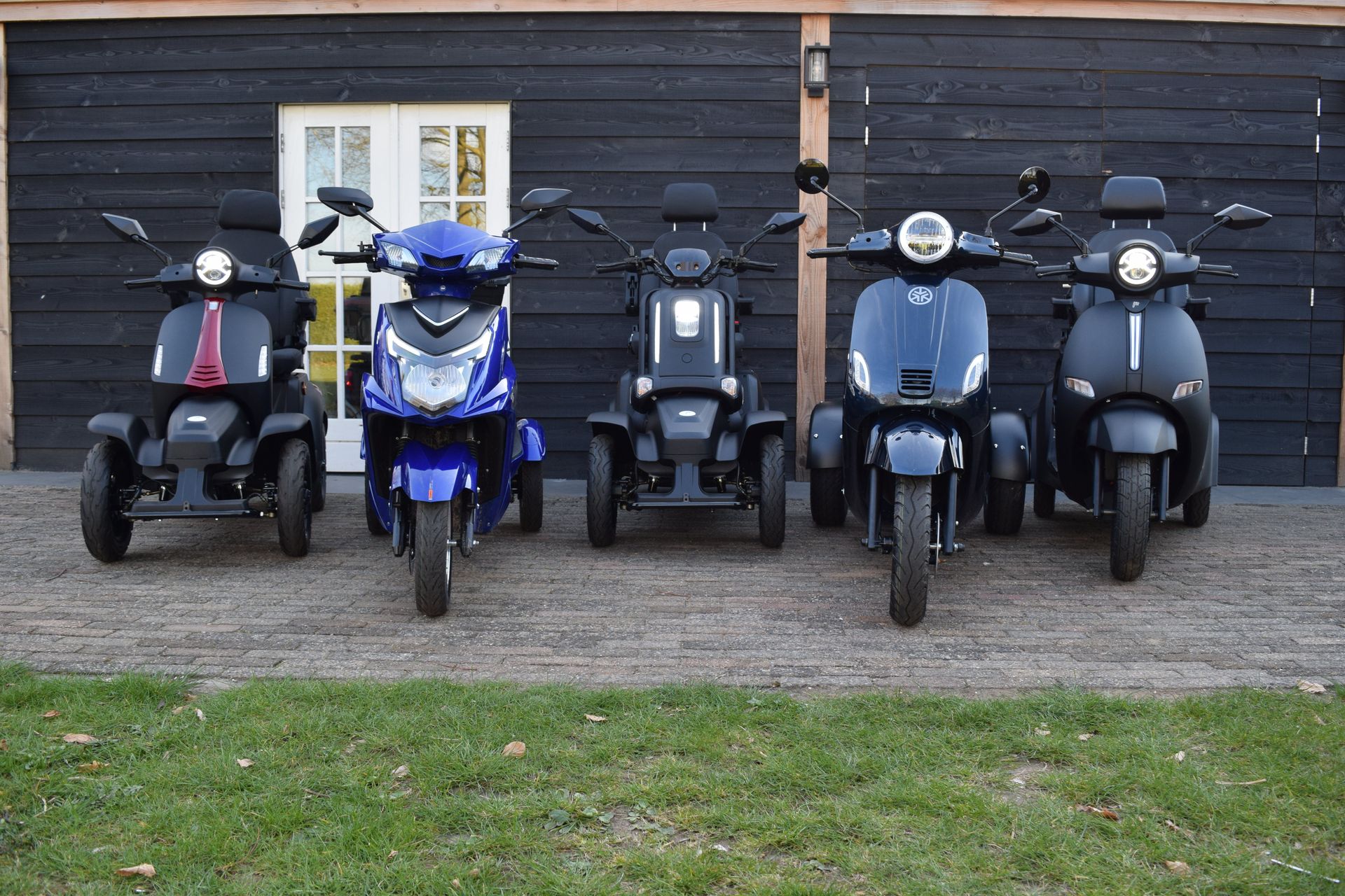 Vijf scootmobielen op een rij, elk met een ander ontwerp en een andere kleur, geparkeerd voor een donker gebouw.