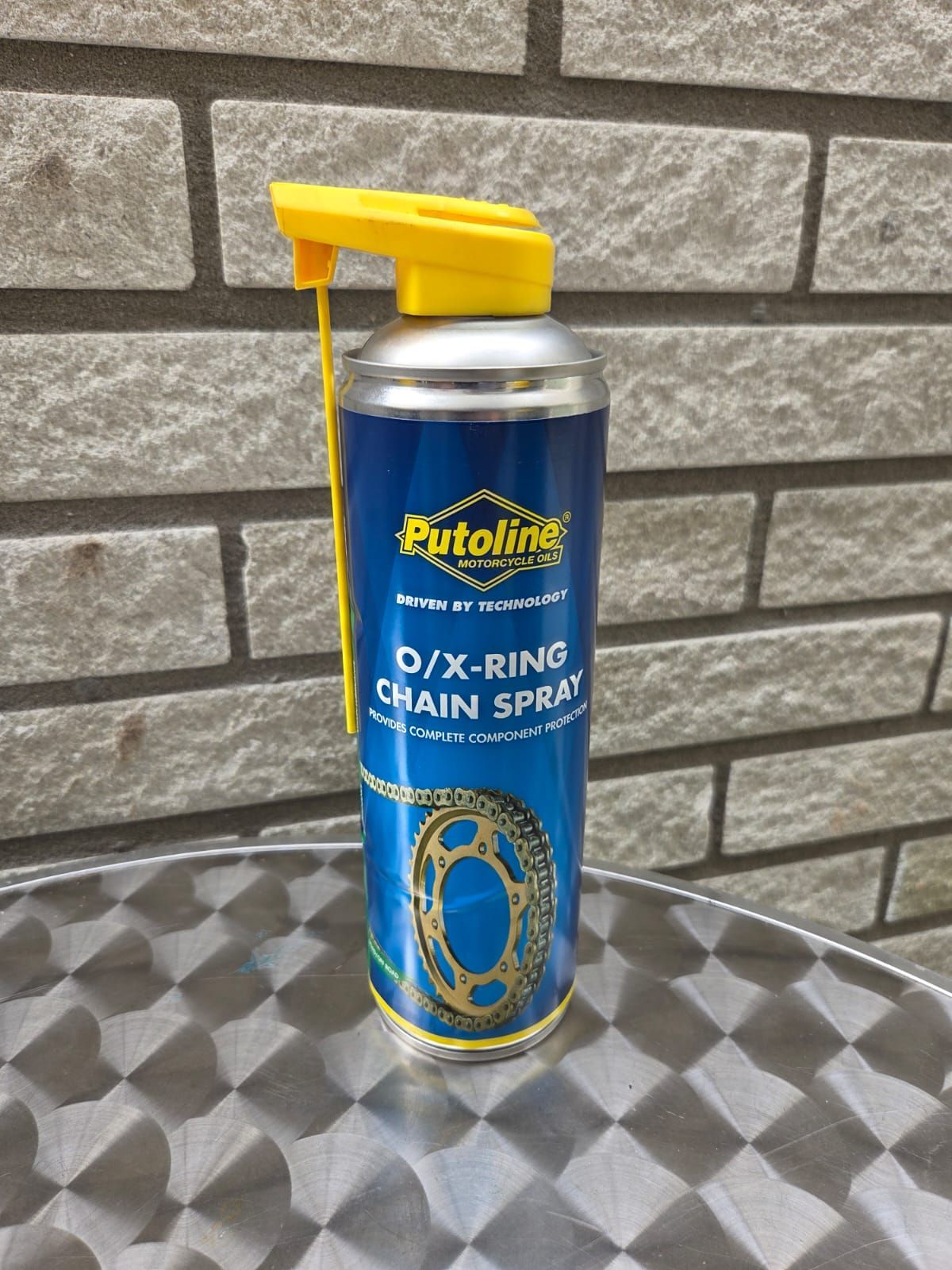 Een bus Putoline O/X-ring kettingspray met een gele spuitmond staat op een tafel met een patroon voor een bakstenen muur.