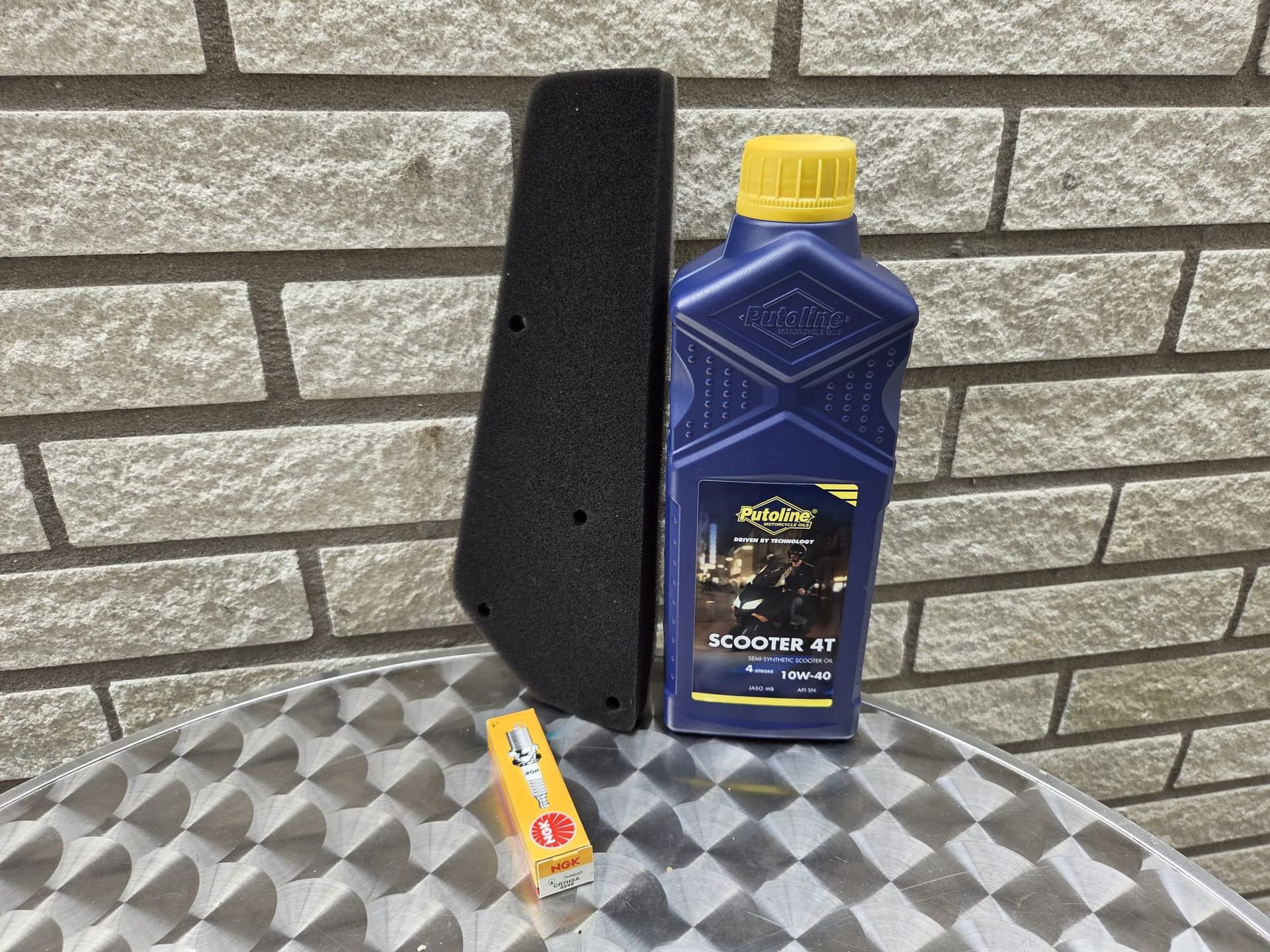 Een zwart luchtfilter, een fles olie en een bougie liggen op een geruite tafel voor een bakstenen muur.