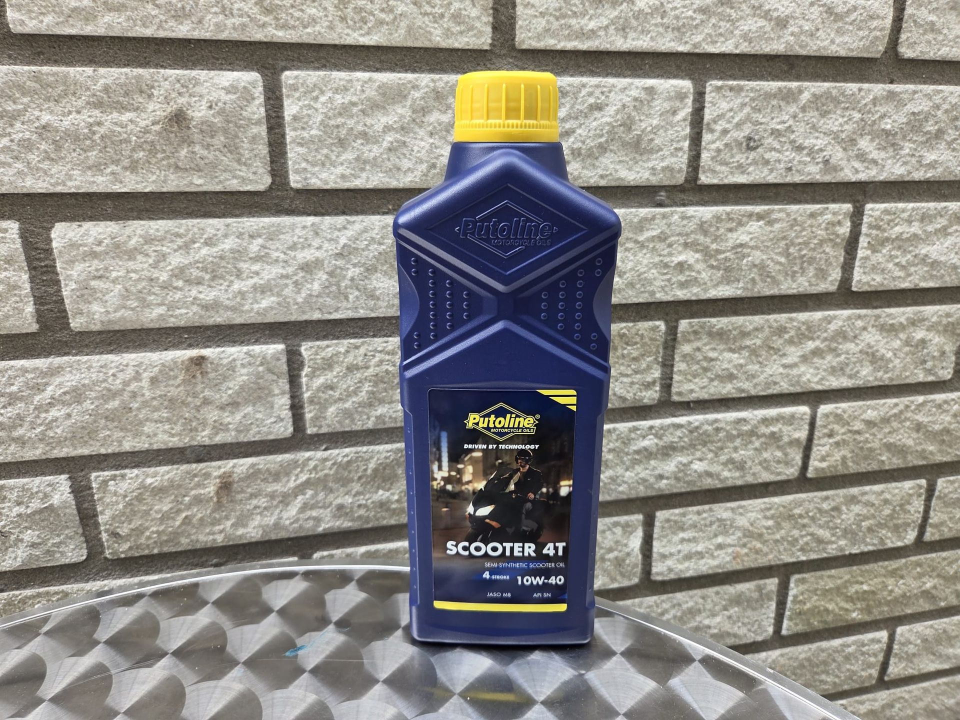 Blauwe fles scooterolie met gele dop, op een metalen ondergrond tegen een bakstenen muur.