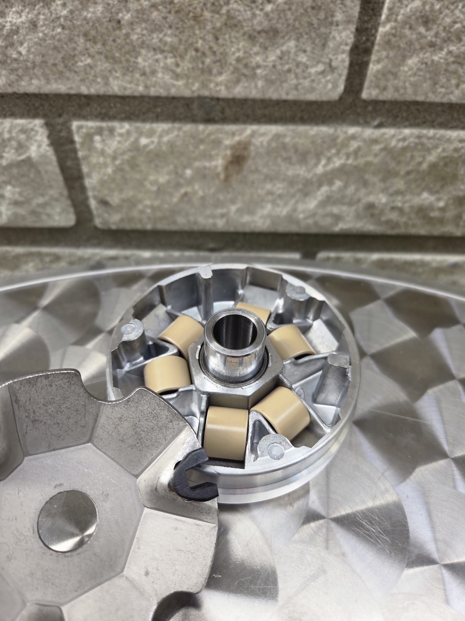 Een scootervariator met beige rollen en een metalen tandwiel op een glanzend oppervlak, tegen een bakstenen muur.