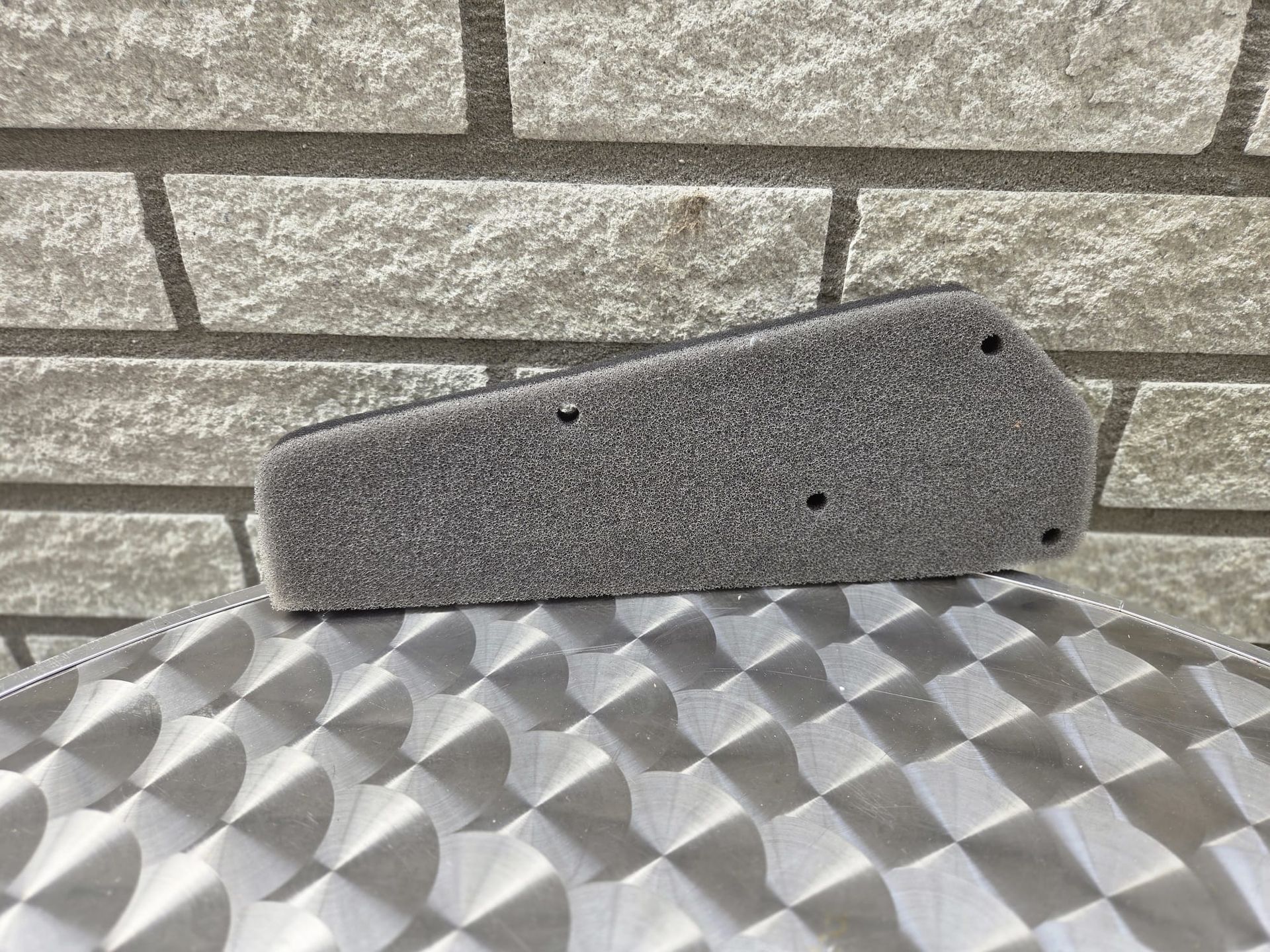 Grijs schuimrubberen luchtfilter op een metalen oppervlak met patroon, tegen een bakstenen muur op de achtergrond.