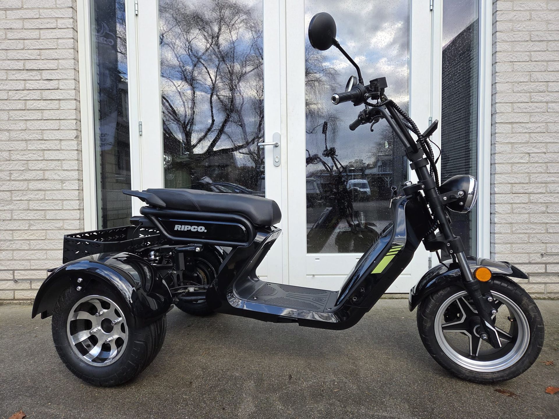 Een zwarte driewielige scooter staat geparkeerd voor glazen deuren; hij heeft een zadel, een stuur en een laadruimte.